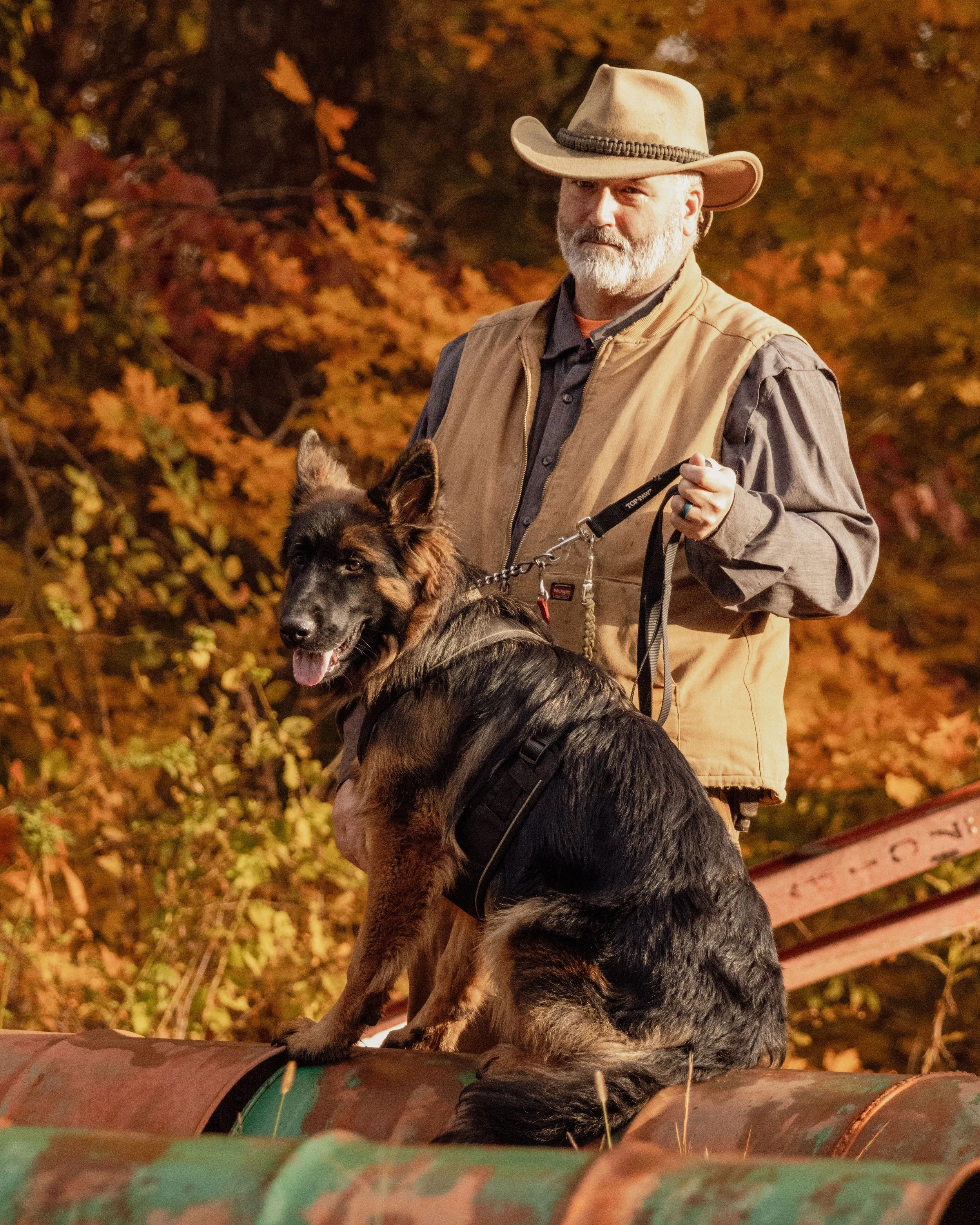 A man in a cowboy hat and tan vest holding a German Shepherd dog on a leash outdoors with autumn leaves in the background.