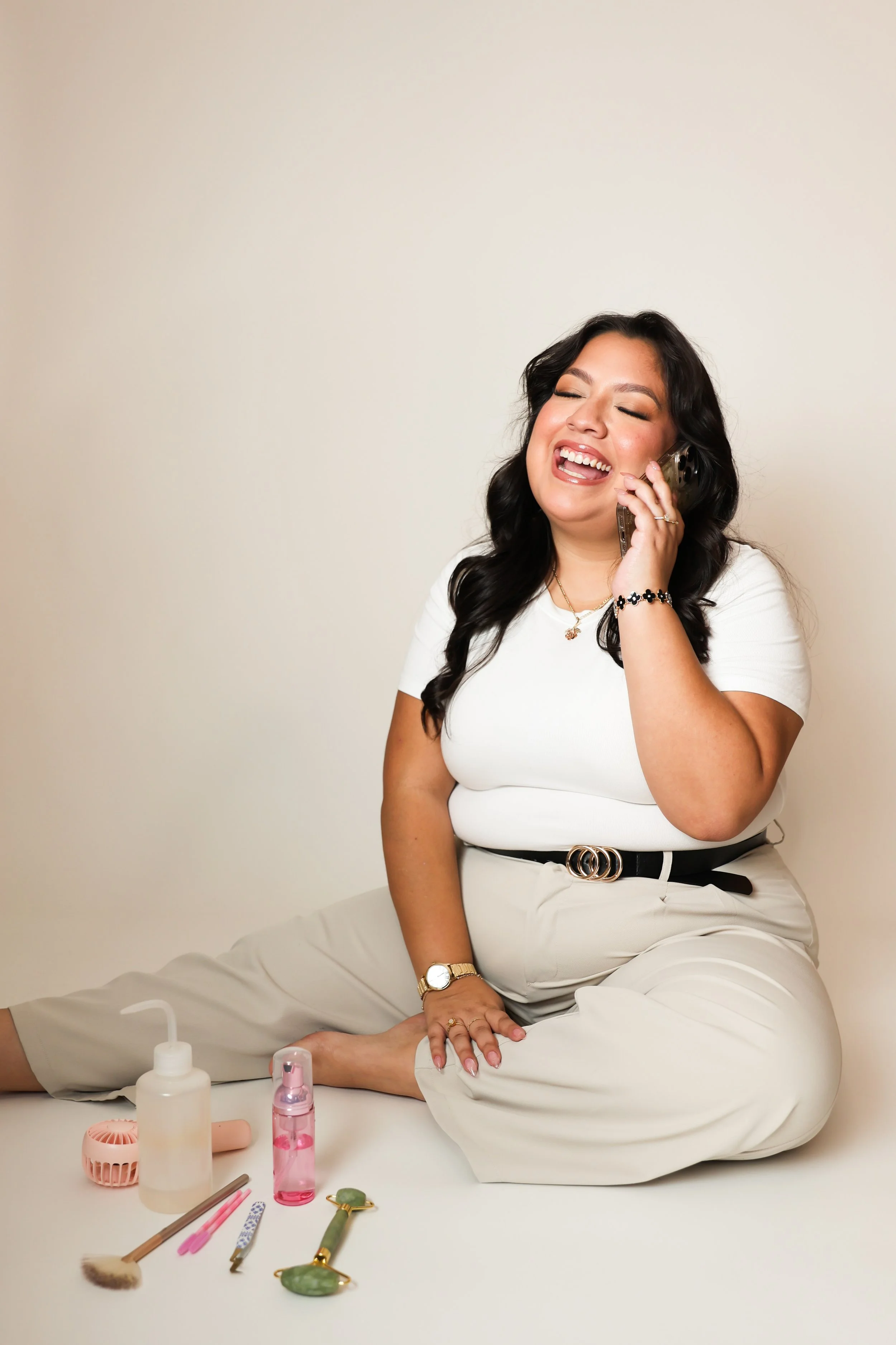 A woman with dark hair, wearing a white shirt and beige pants, sitting on the floor, smiling and talking on her phone with beauty tools and skincare products around her.