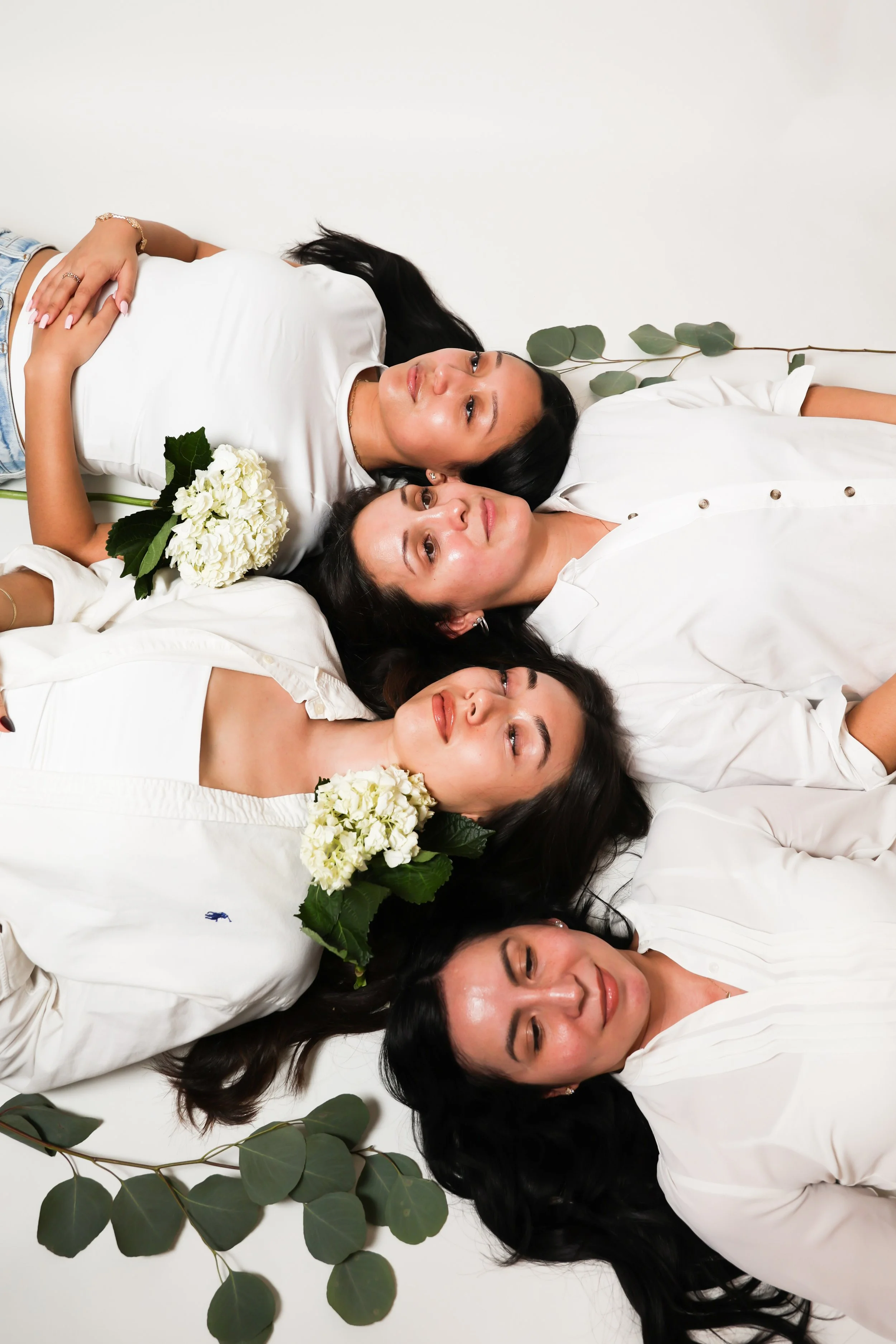 Five women lying on their backs on a white floor with green leaves around them, two of them holding white hydrangea flowers.
