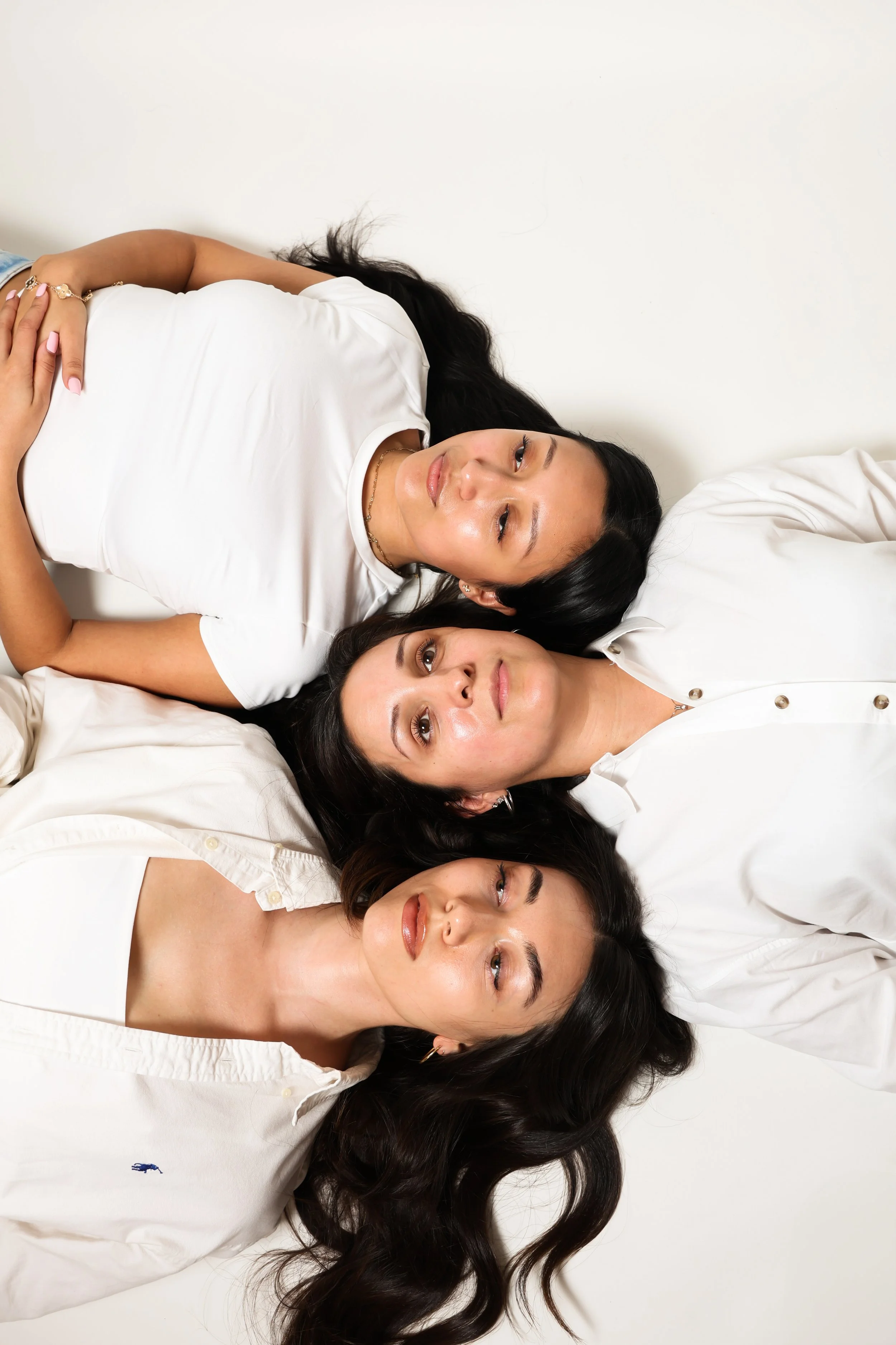 Three women lying on the floor with their heads close together, facing up against a white background.