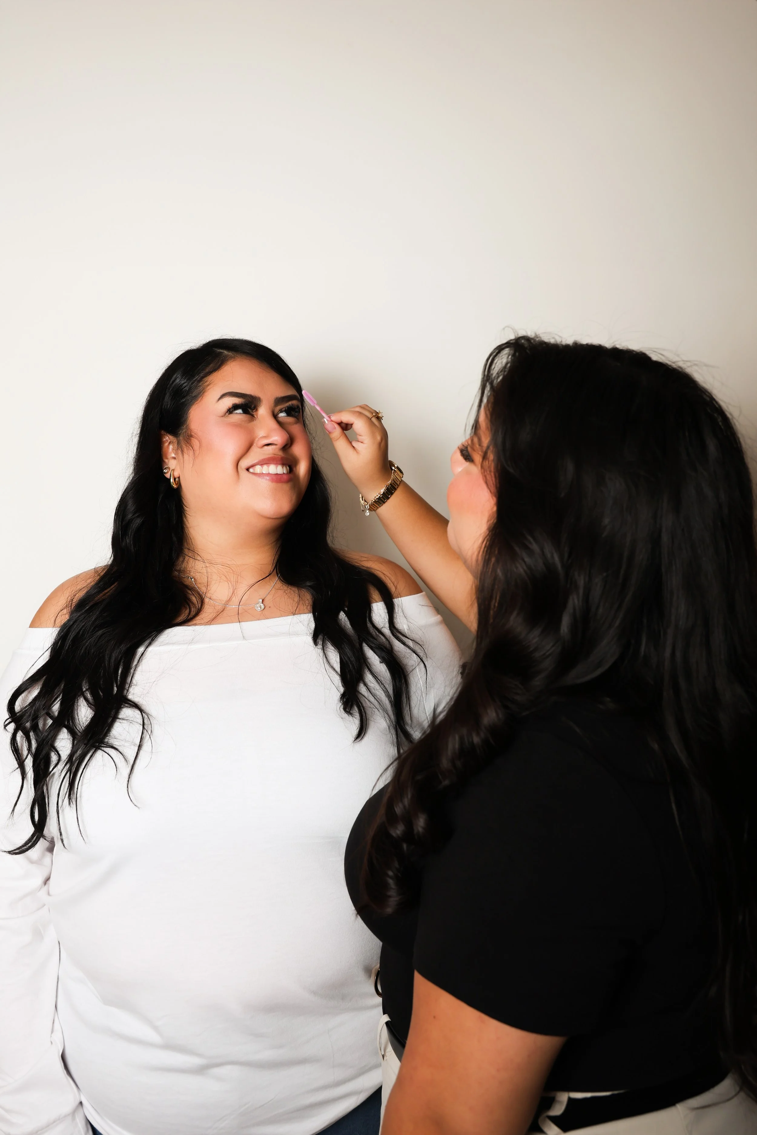 A woman with long black hair smiling as another woman applies makeup on her face, holding a makeup brush close to her eyebrow.