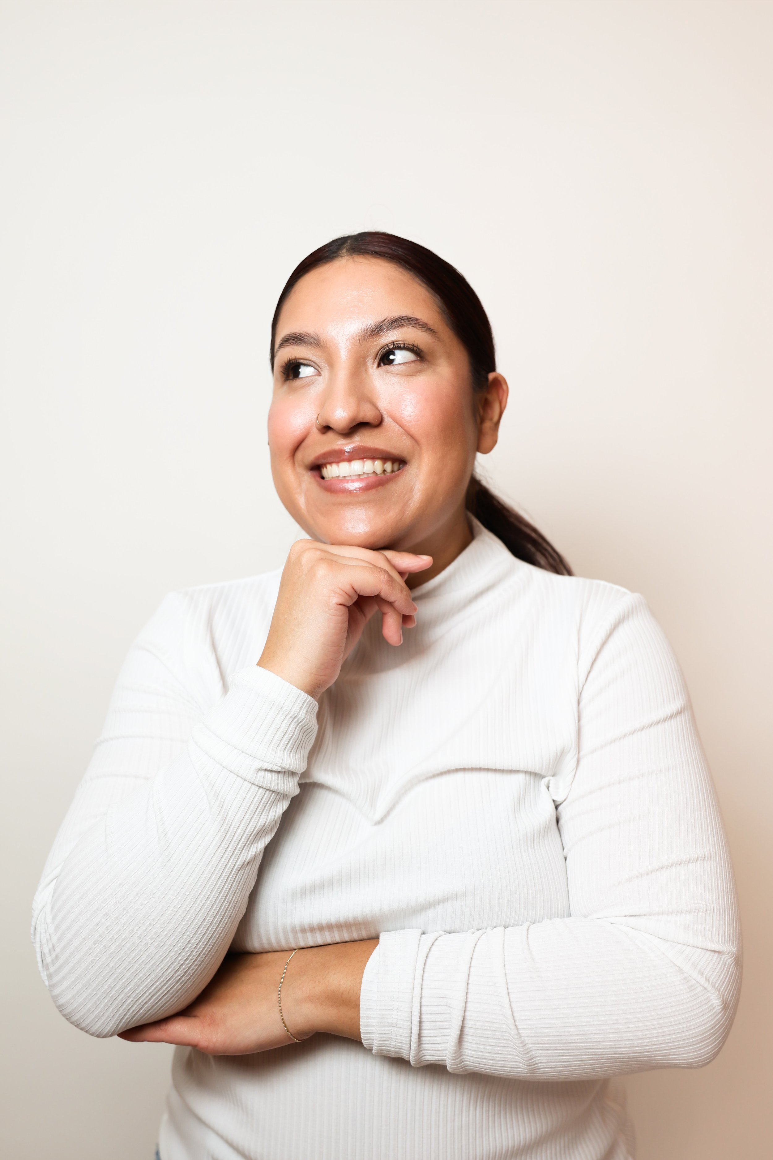 A woman with dark hair tied back, smiling with her hand resting under her chin, wearing a white long-sleeve shirt, standing against a plain white background.
