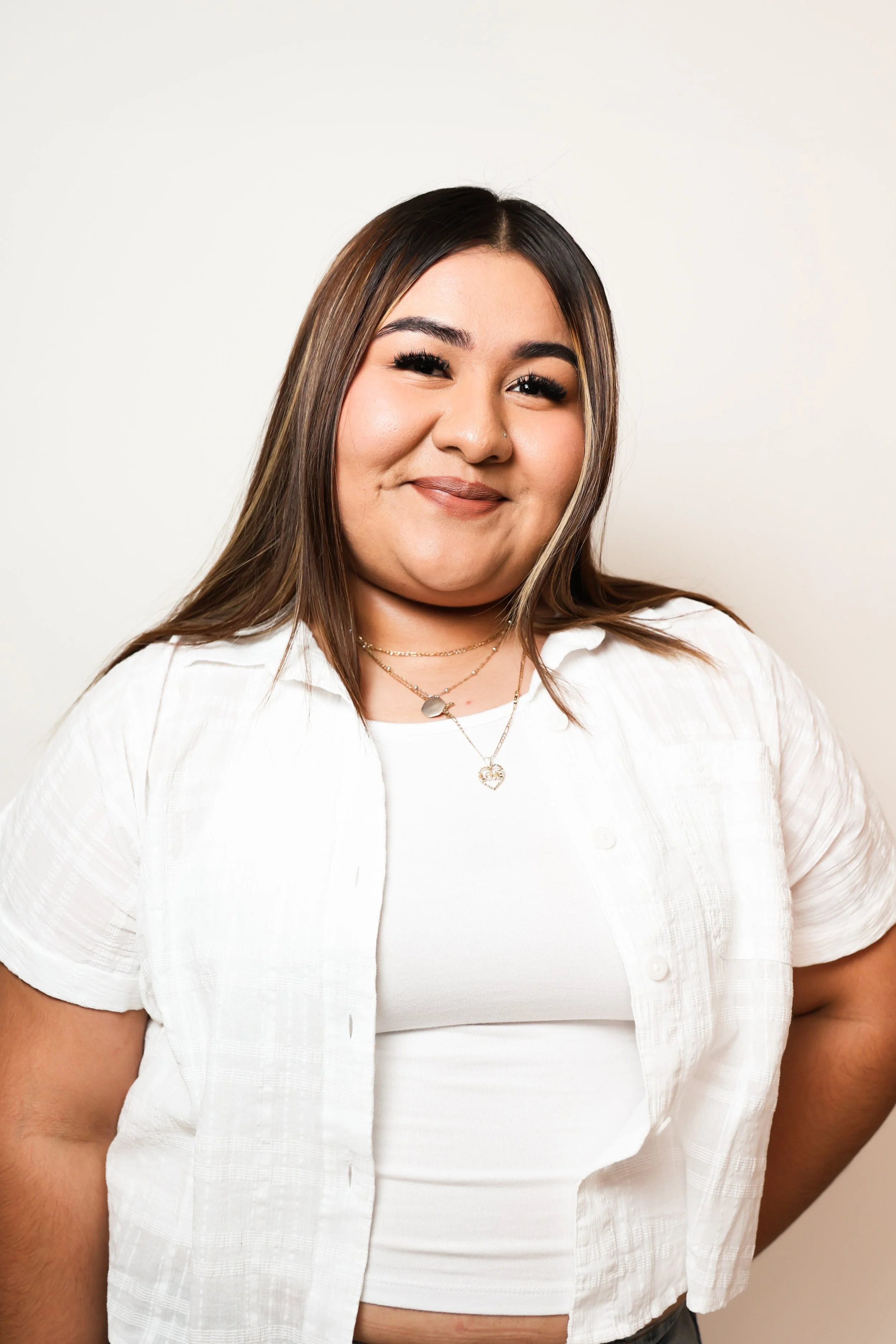 A young woman with dark brown hair, wearing a white top and layered necklaces, smiling at the camera against a plain white background.