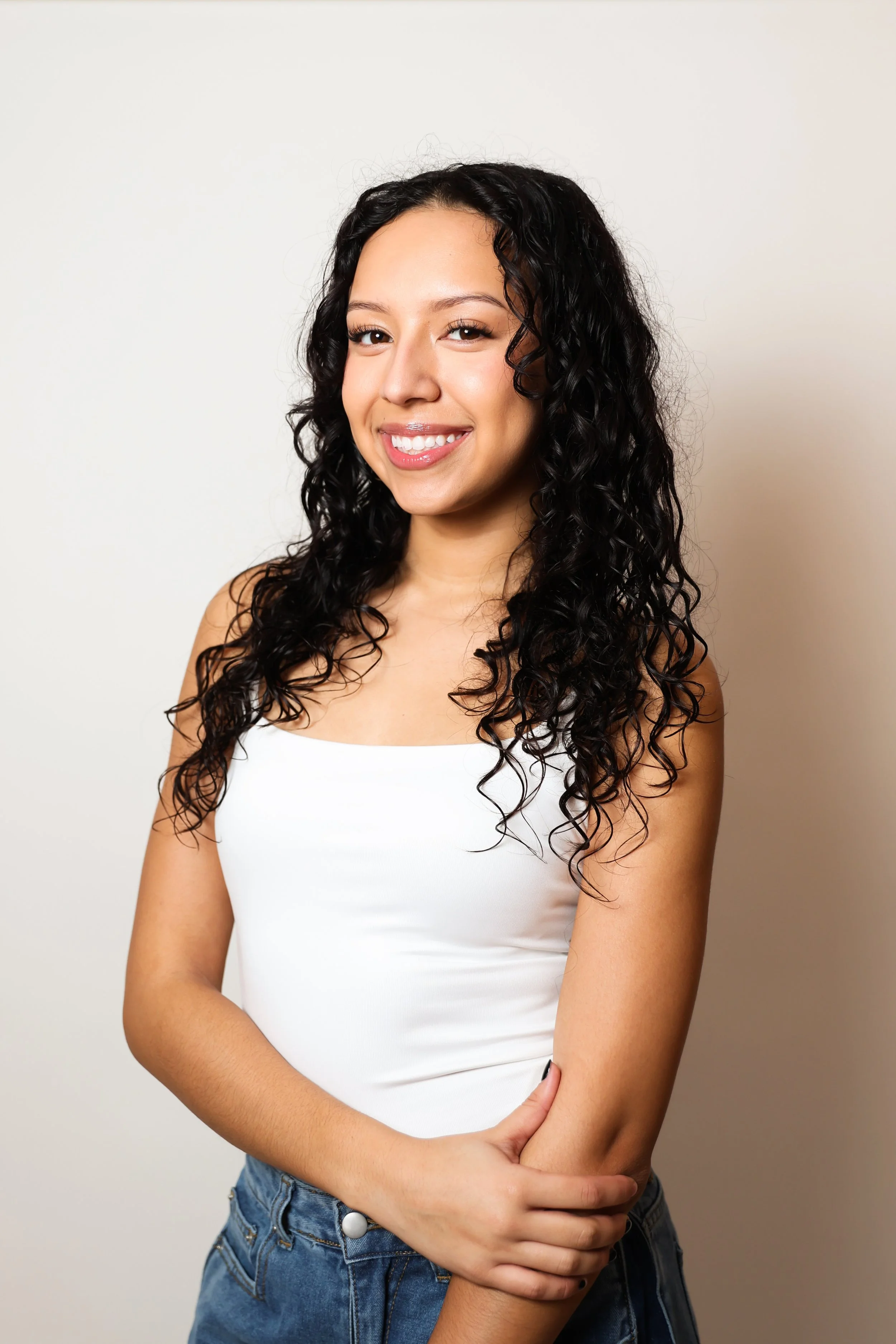 A young woman with curly black hair smiling and wearing a white tank top and blue jeans, standing against a plain off-white background.