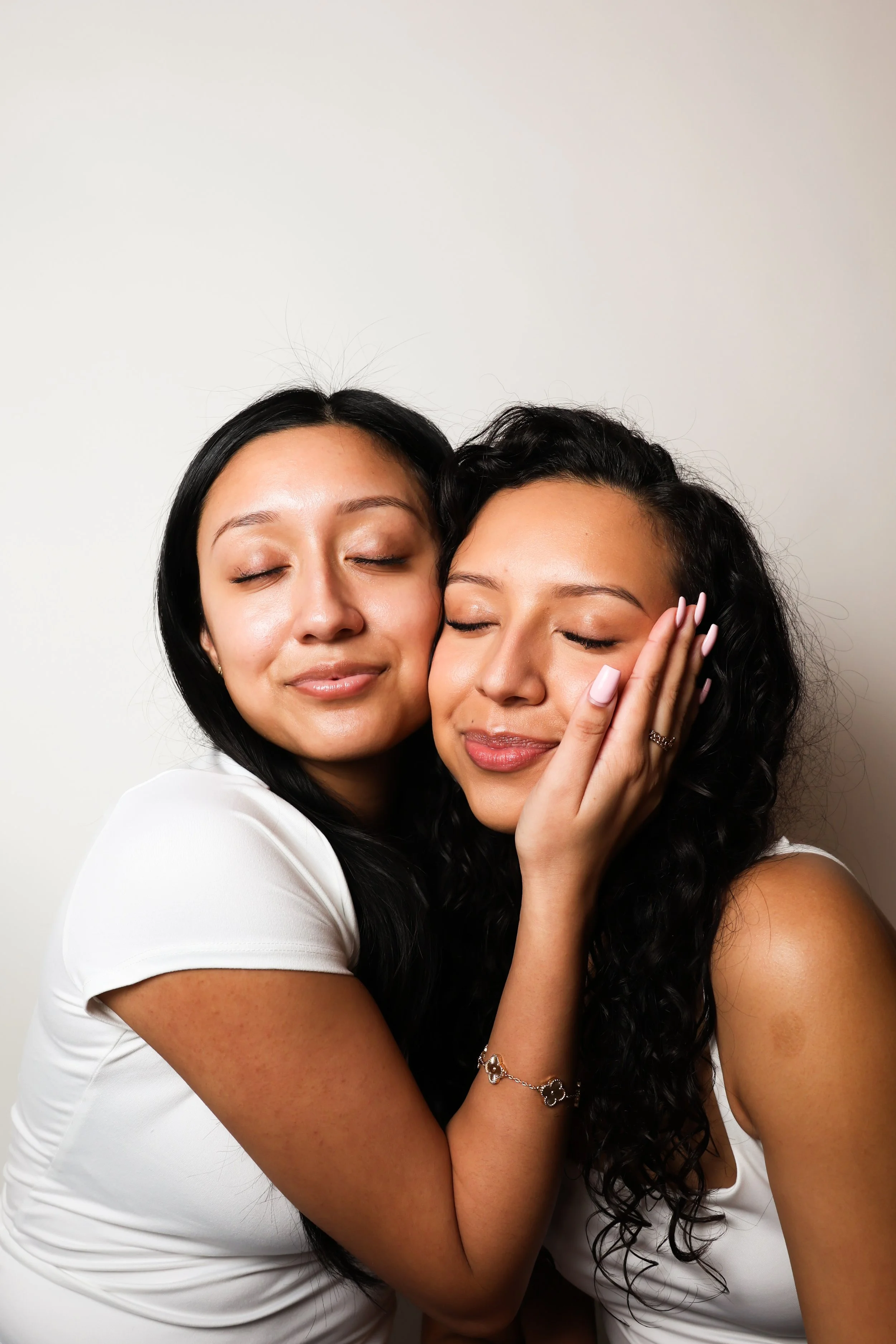 Two women with closed eyes hugging affectionately against a plain white background.
