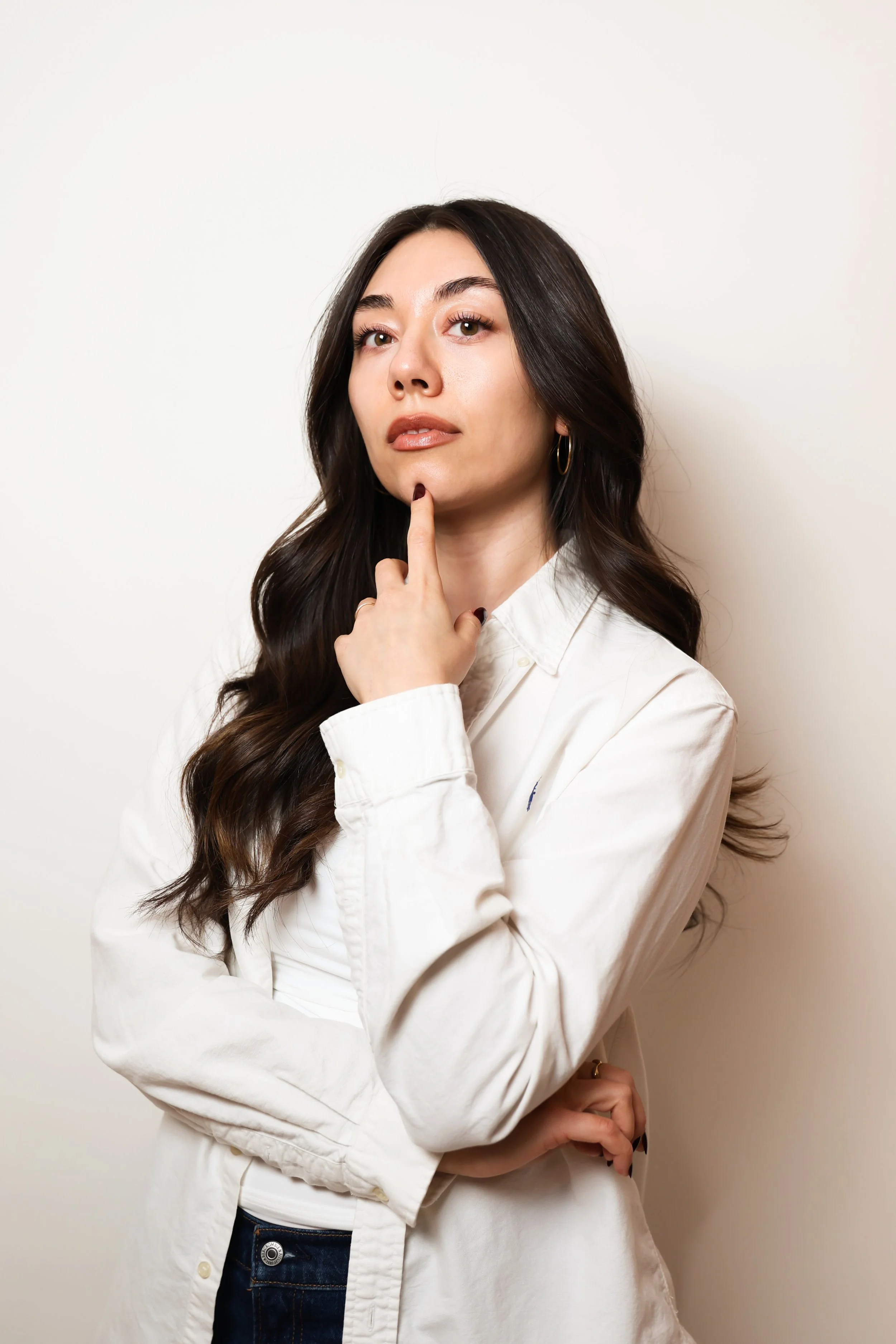 A woman with long dark hair, wearing a white shirt, is touching her chin with her index finger, looking thoughtfully at the camera against a plain white background.