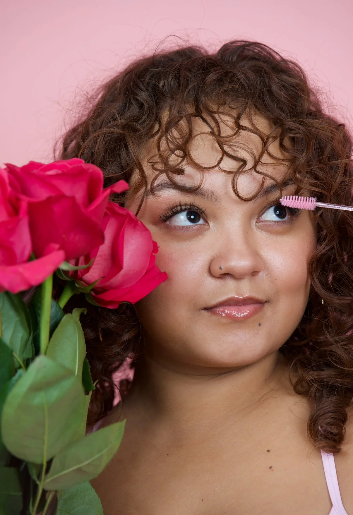 A woman with curly hair and light makeup is looking to her right while holding pink roses near her face. A pink background and a mascara wand are visible.