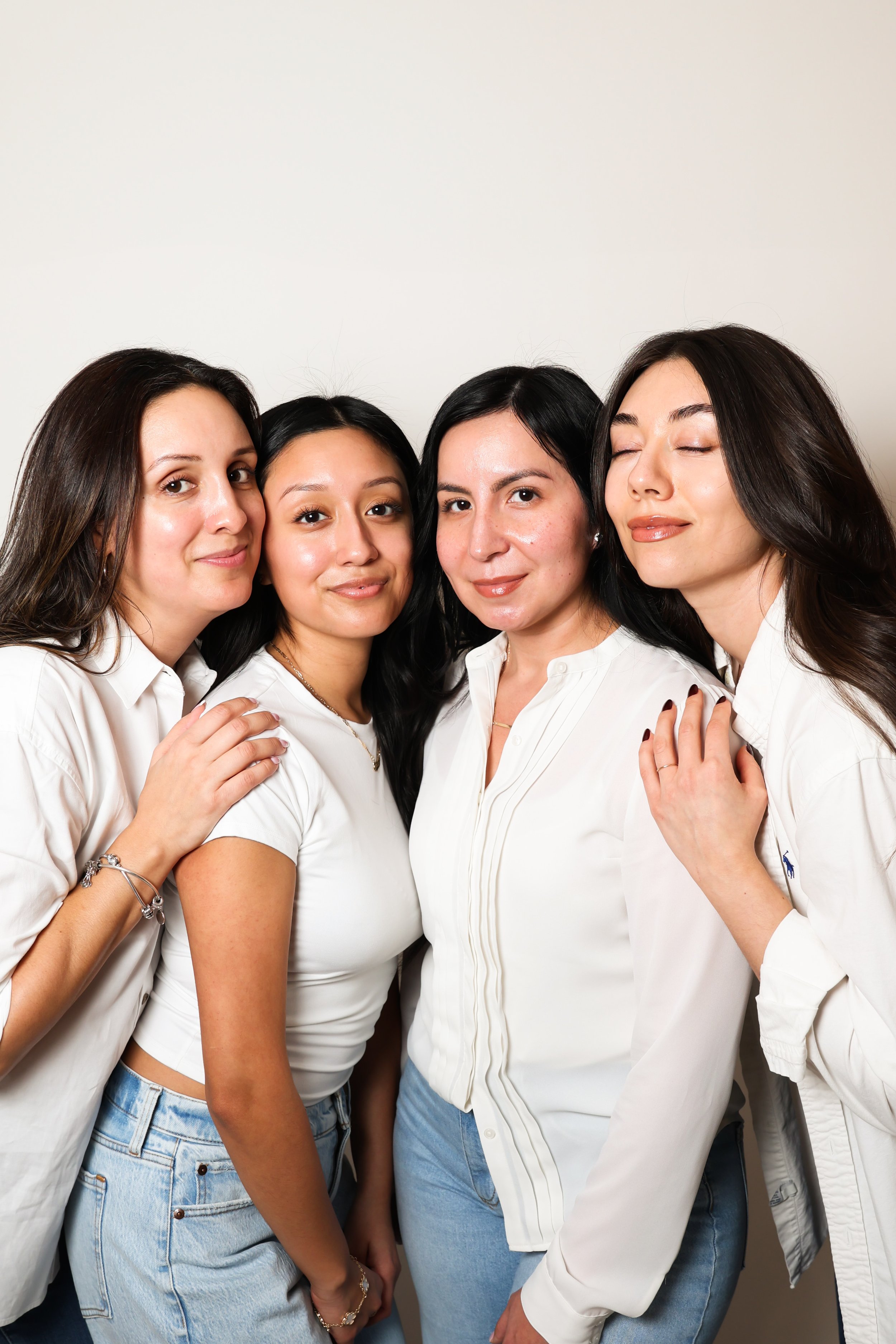 Four women standing close together, smiling and embracing each other against a plain white background.