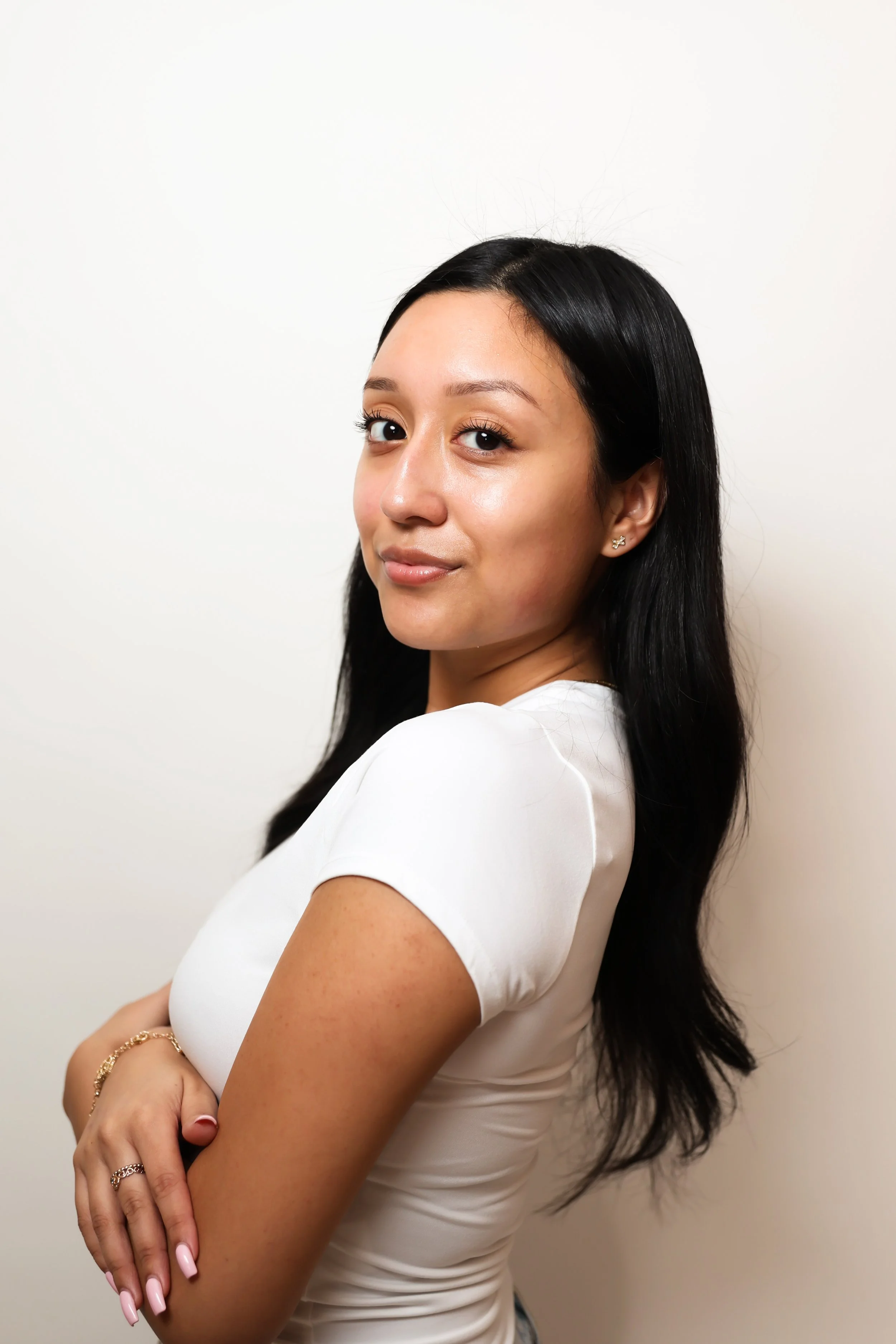 A young woman with long black hair and light makeup standing against a plain white background, wearing a white top with short sleeves and gold jewelry, looking at the camera with a slight smile.