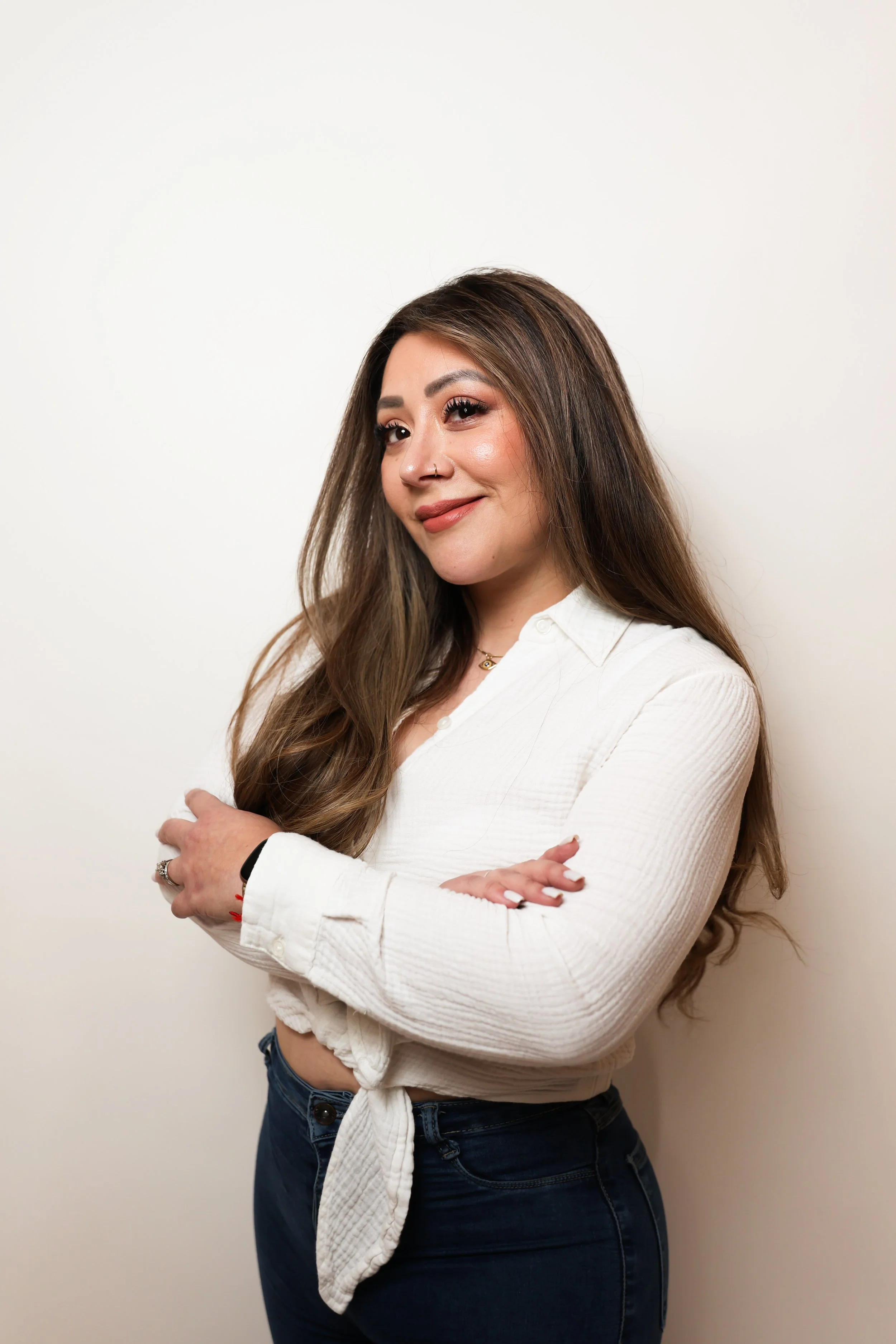 A young woman with long brown hair wearing a white shirt and dark jeans standing against a plain white background.