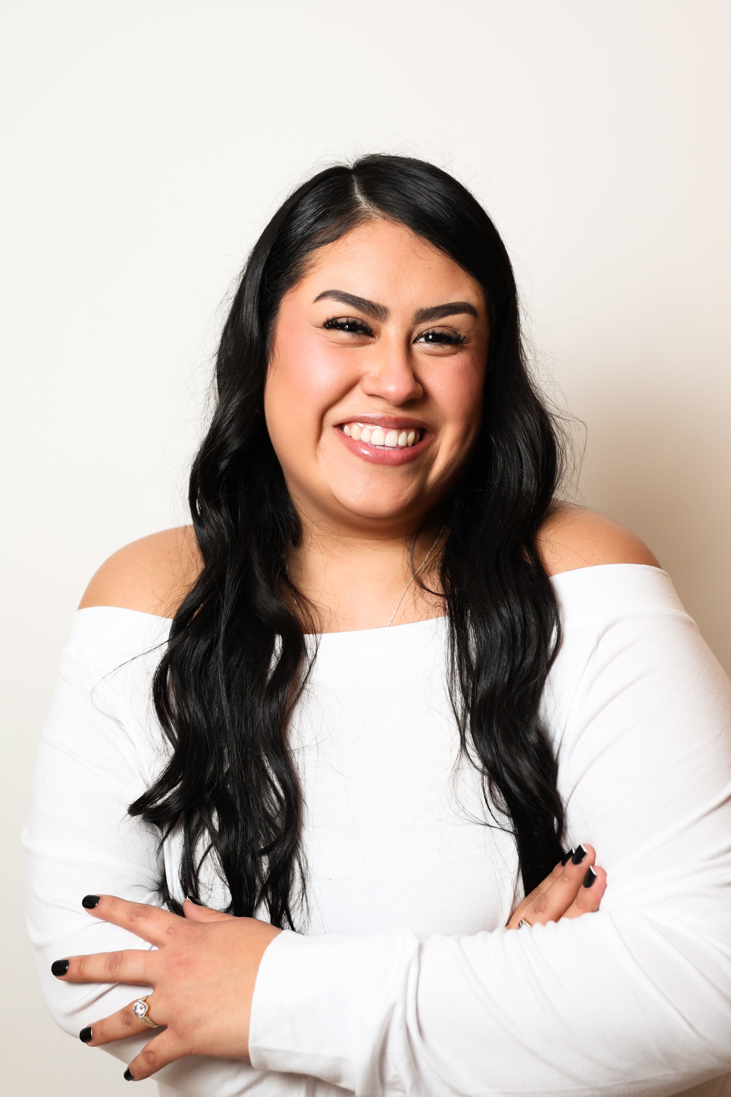 A smiling woman with long black hair, wearing a white off-shoulder top, standing against a plain white background.