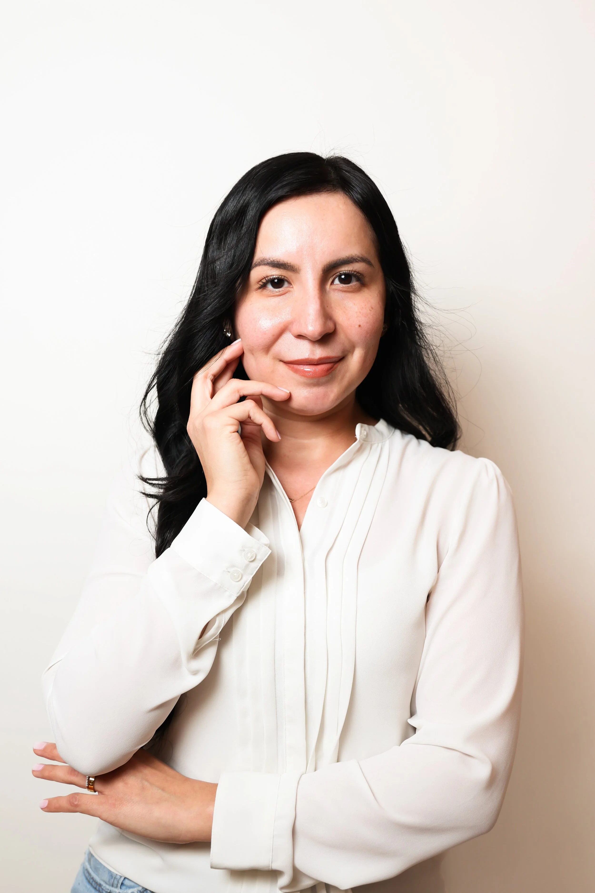 A woman with long black hair, wearing a white blouse, standing against a plain white wall, posing with her hand near her face and smiling softly.