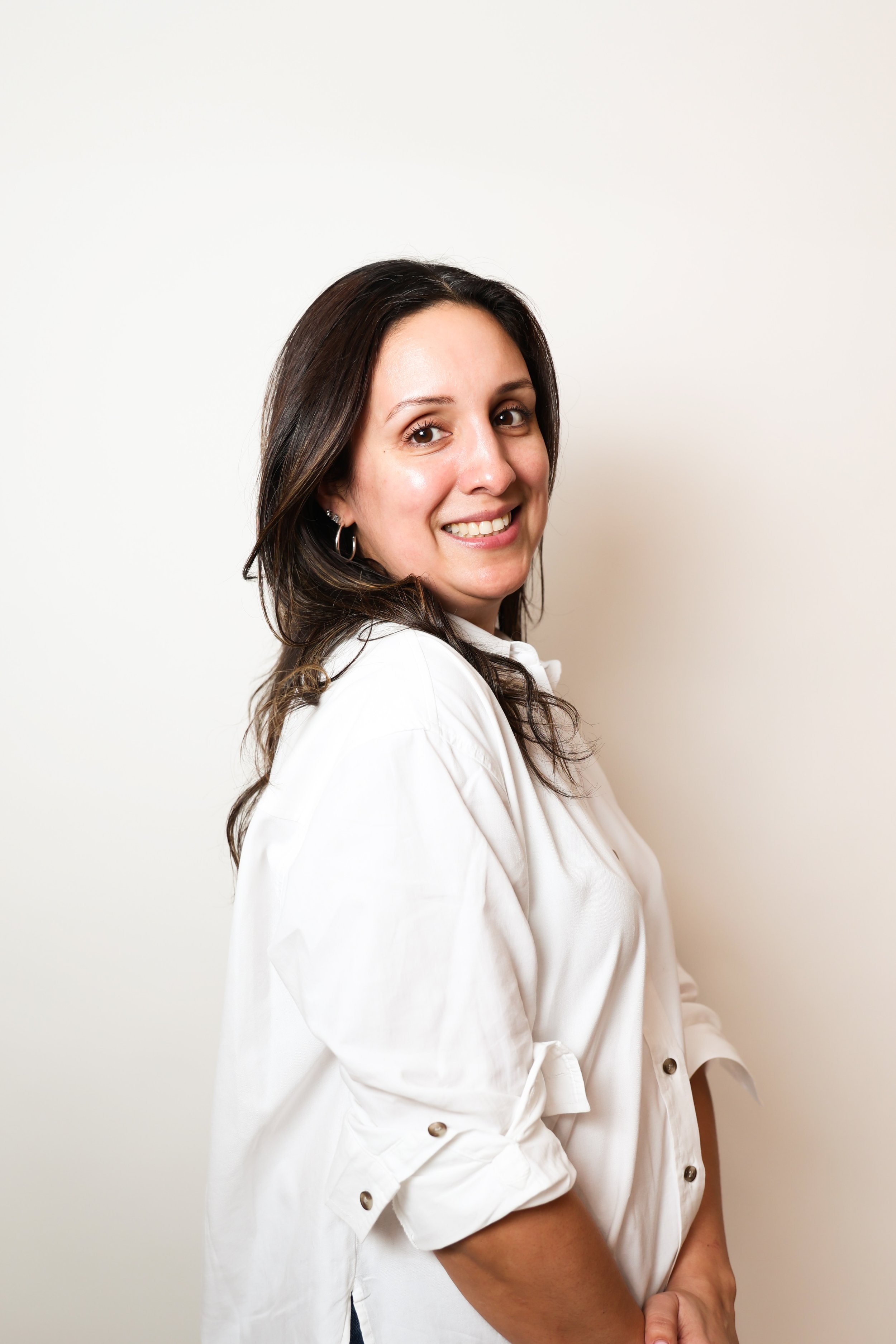 A woman with shoulder-length dark brown hair smiling, wearing a white button-up shirt with rolled-up sleeves, standing against a plain white background.