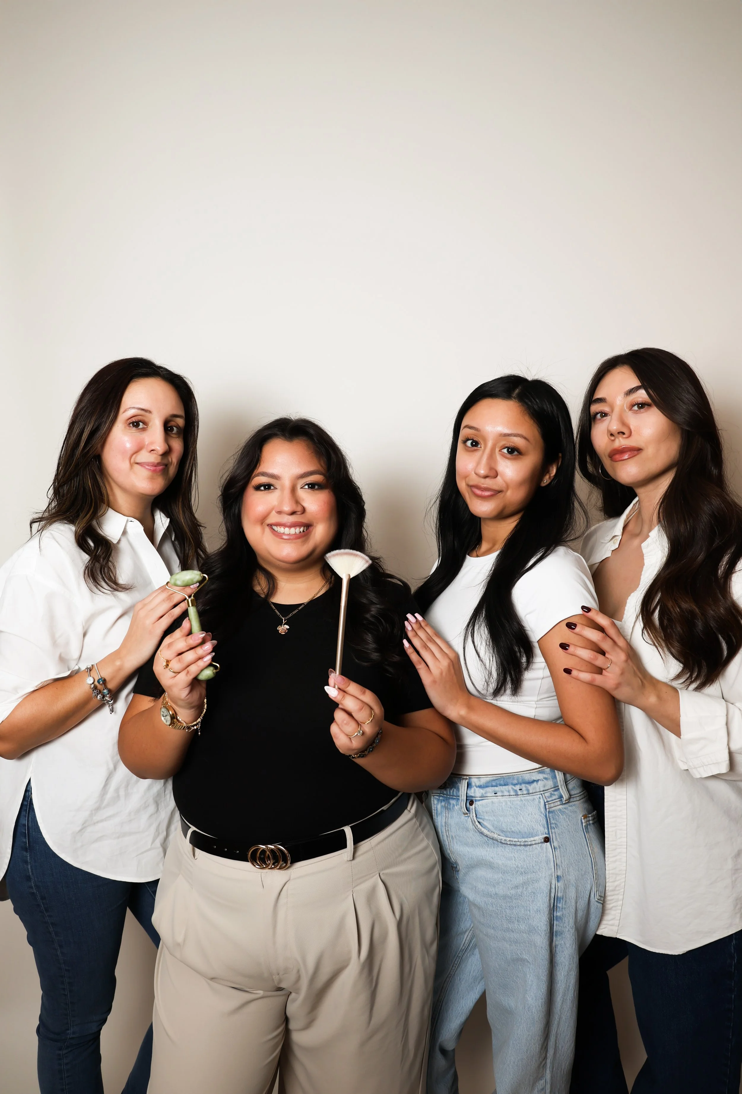 Group of four women posing together, smiling, against a plain background, with beauty or skincare tools and accessories.
