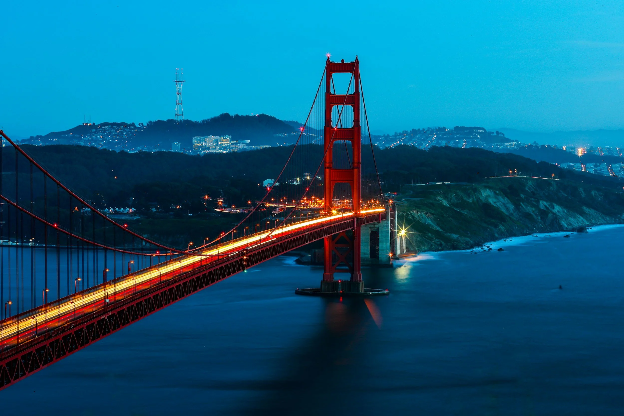 Night view of the Golden Gate Bridge in San Francisco, California, with city lights and hills in the background, and the water below.