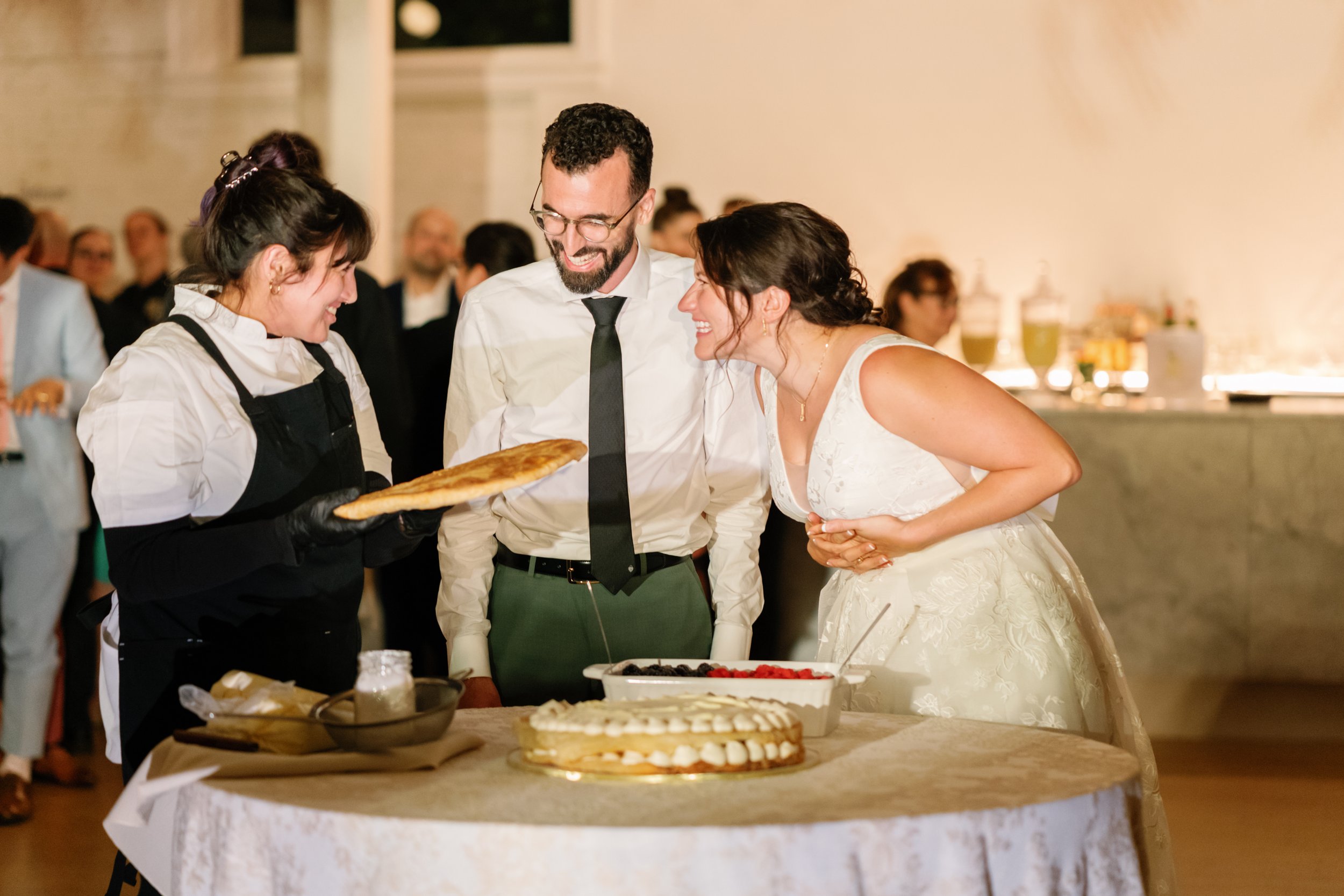 A bride and groom at their wedding cake cutting celebration, interacting with a waitress holding a pizza at the reception.
