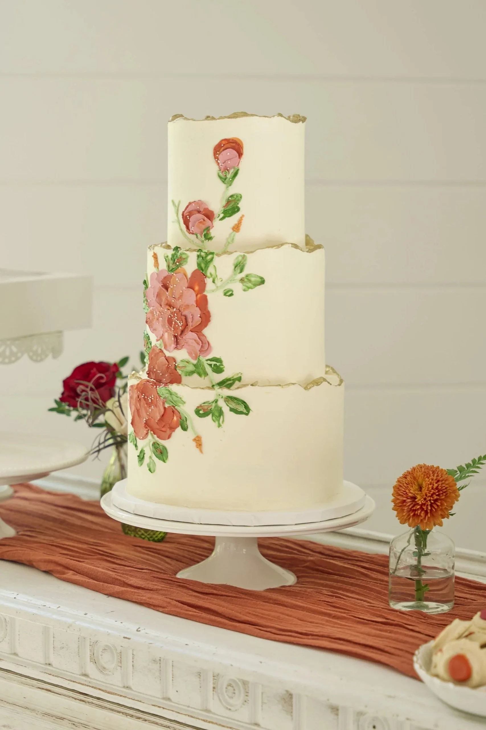 A three-tiered white wedding cake with floral decorations on each tier, placed on a cake stand on a wooden table, with small flowers in a glass vase nearby.