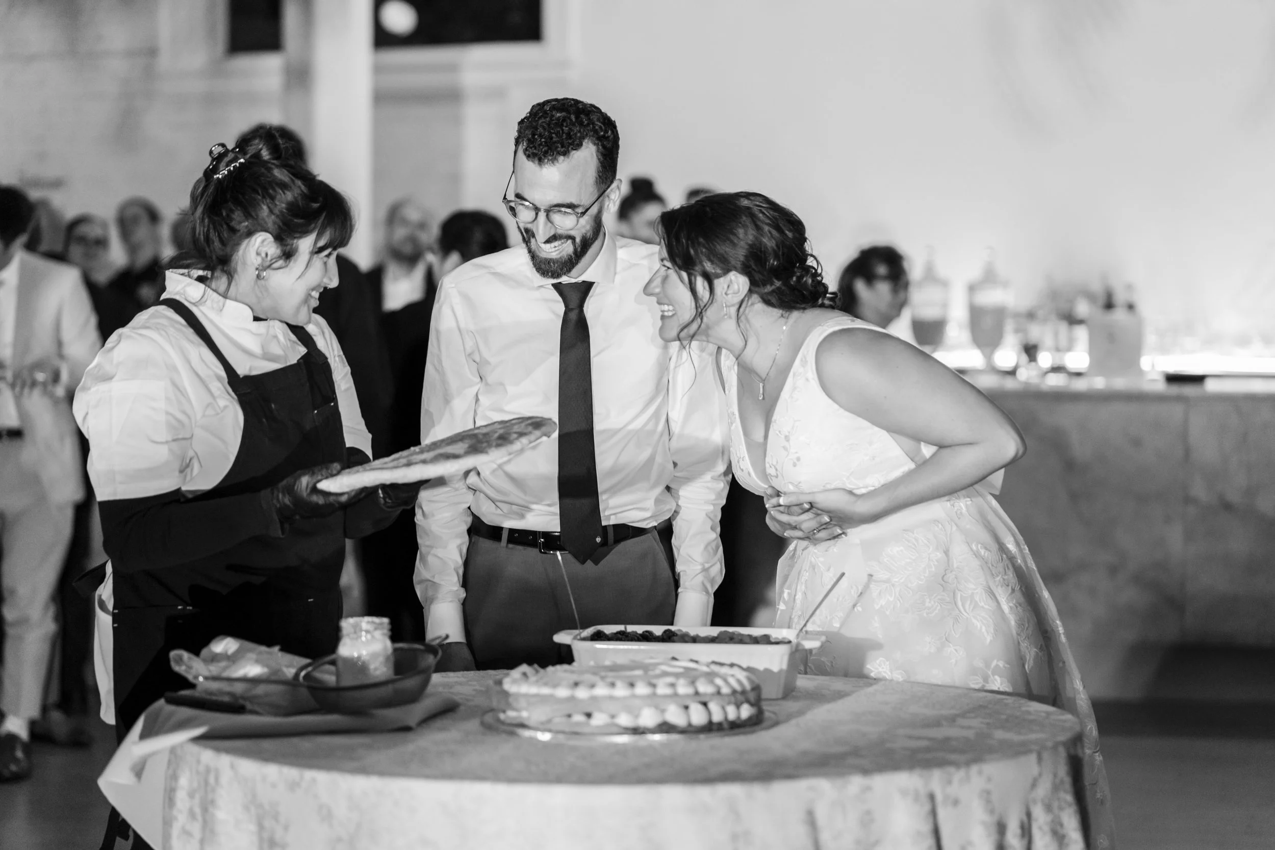 Bride and groom smile and laugh as they look at a server holding a dish, standing at a wedding reception table with cake and food. Guests are in the background.