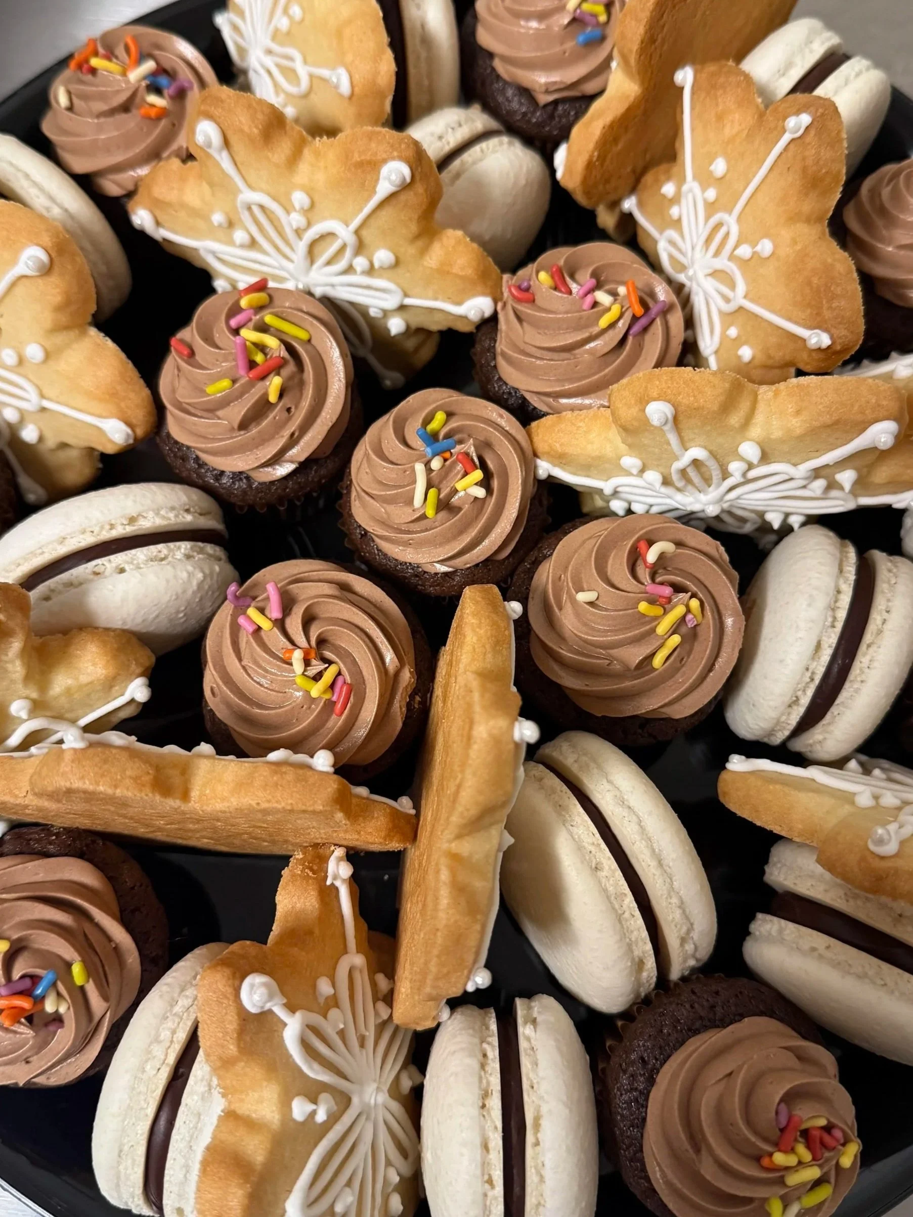 Assorted cookies and cupcakes decorated with white icing, chocolate frosting, and colorful sprinkles on a black platter.