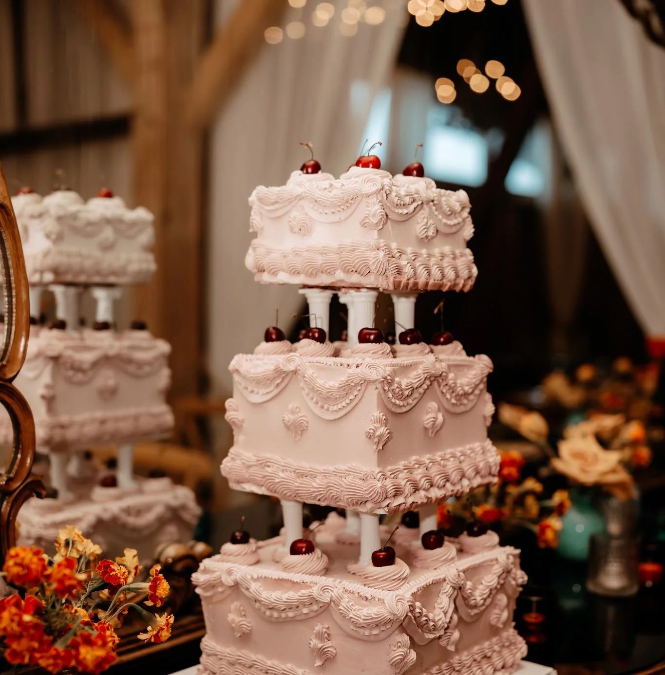 Three-tiered pink wedding cake decorated with cherries on top, with ornate piping and draped icing, set in a rustic barn setting.