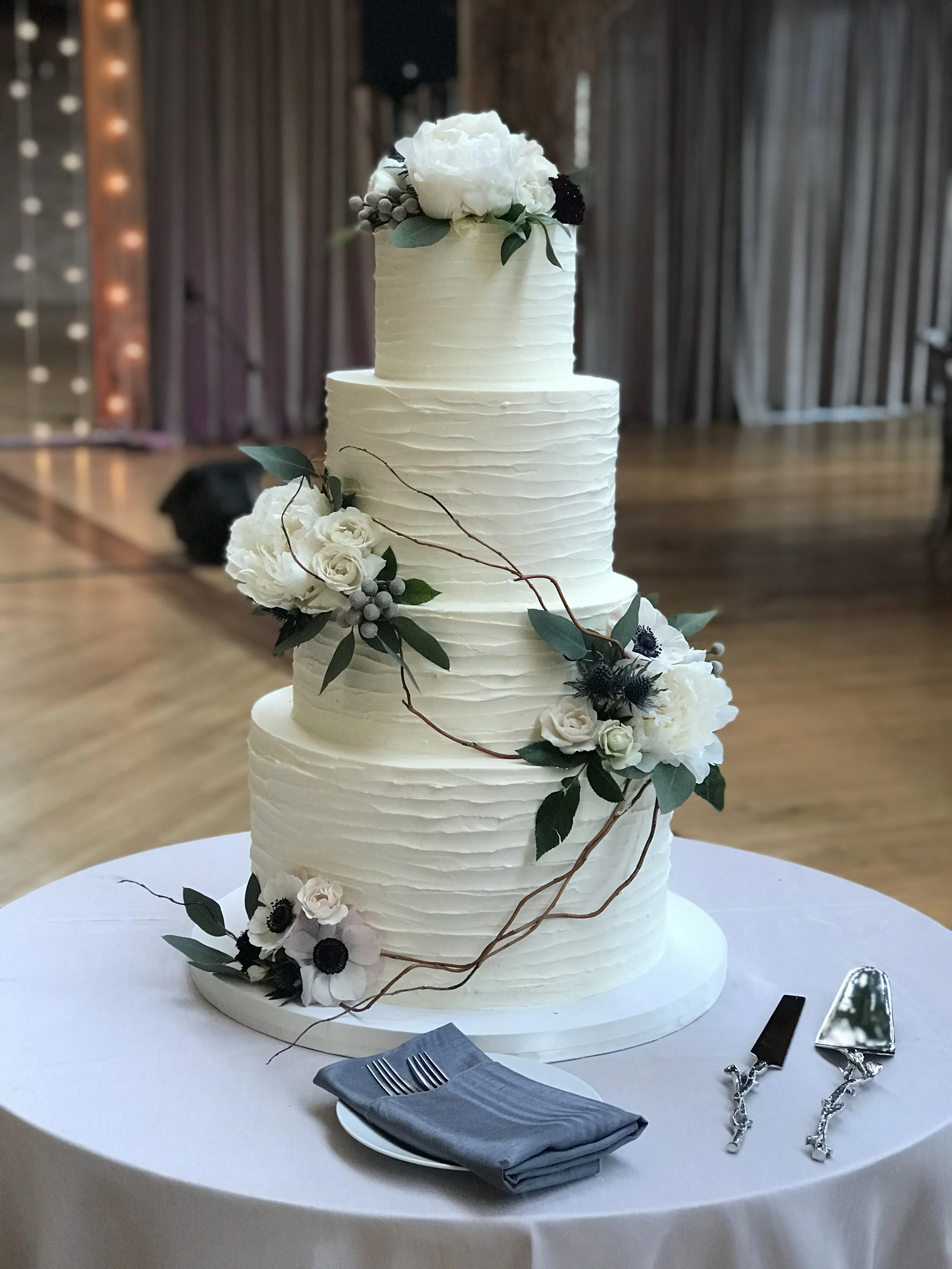 Three-tier white wedding cake decorated with white and dark flowers, green leaves, and a vine, placed on a round table with a gray napkin, forks, and cake server in front of it.