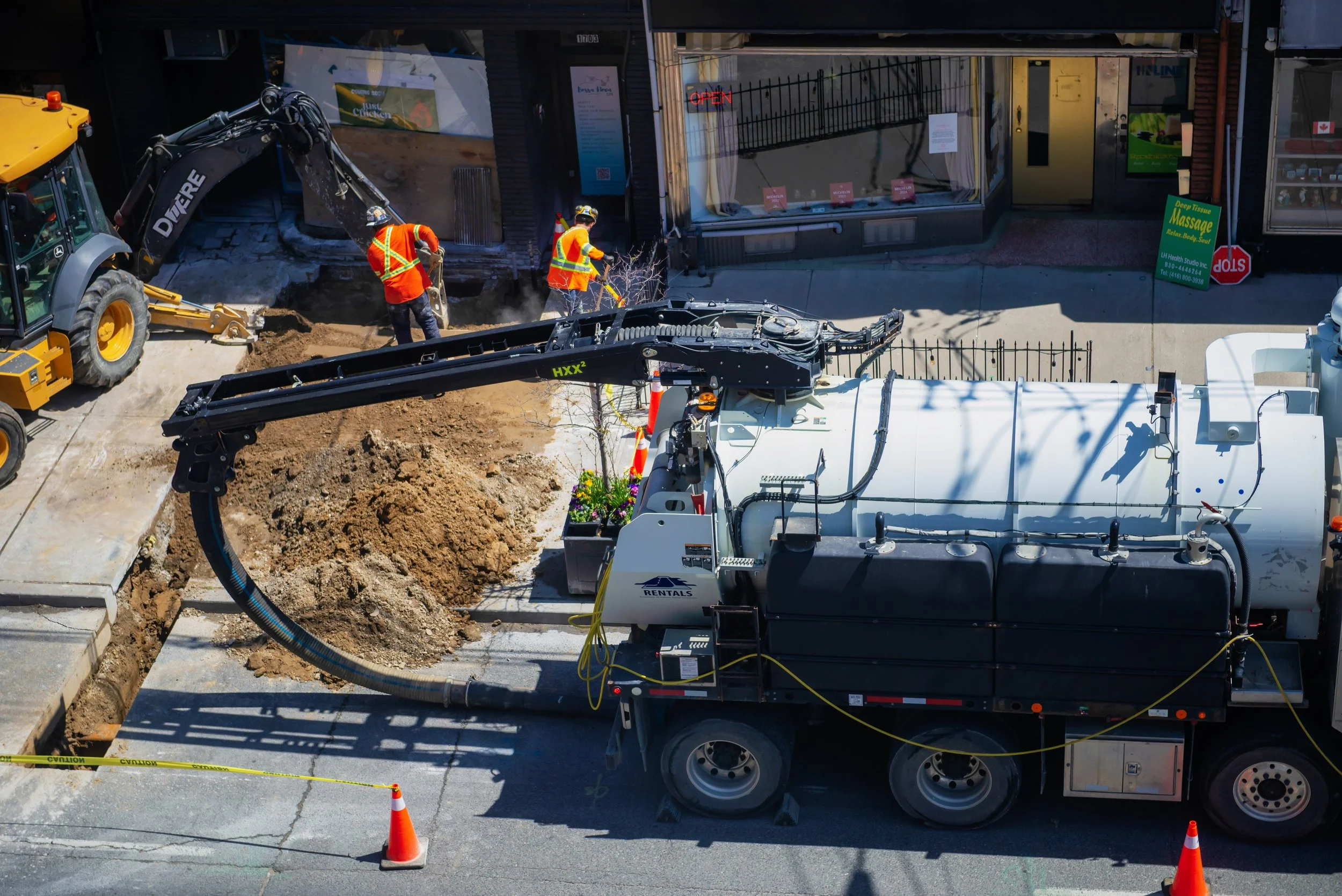 Construction workers in orange safety vests and helmets working on a trench on the street, with a large vacuum excavation truck and a backhoe loader nearby.