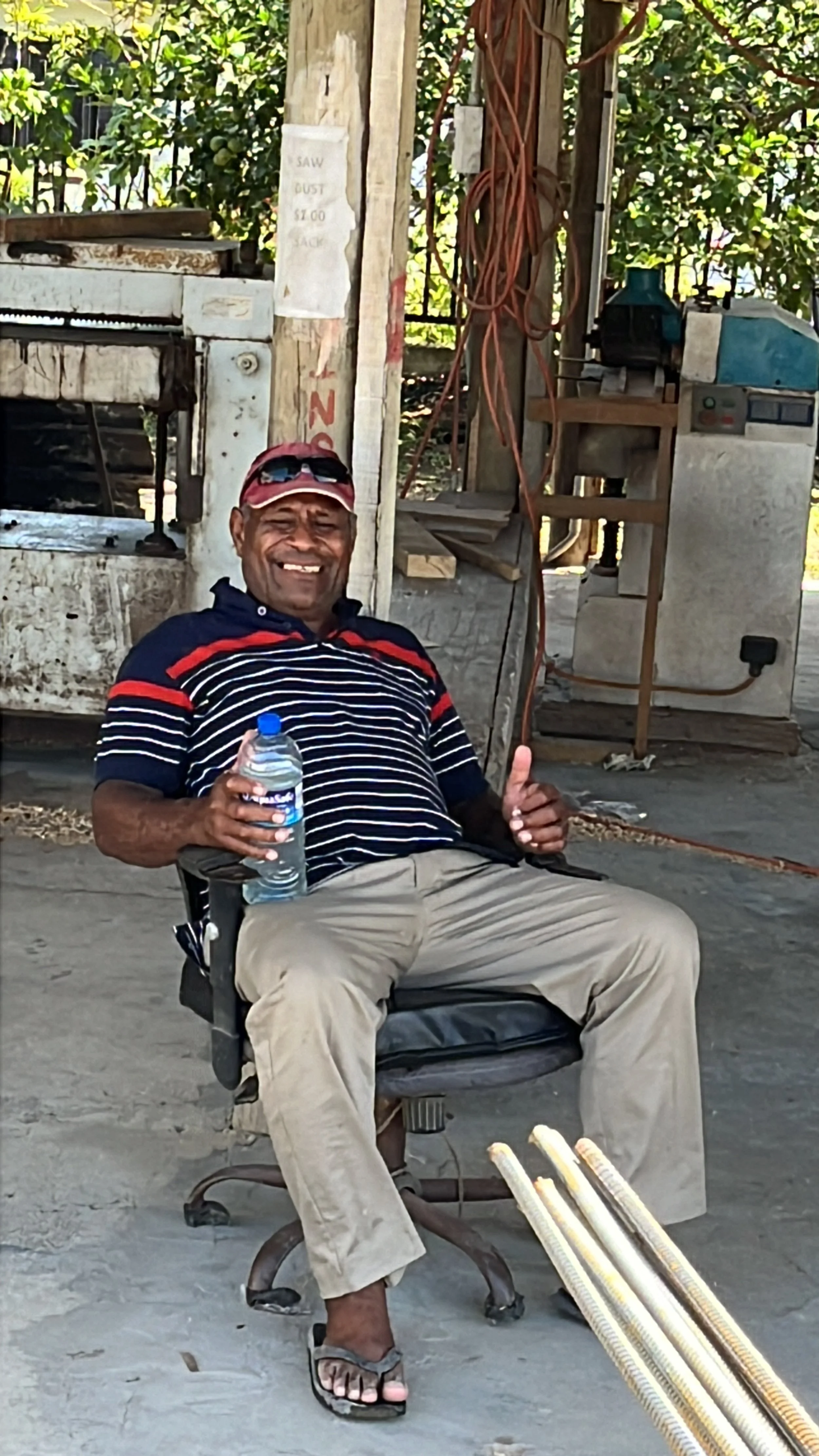A man sitting on an office chair outdoors, smiling and holding a bottle of water, wearing a striped polo shirt, khaki pants, flip-flops, and a red cap, with construction pipes on the ground in front of him.