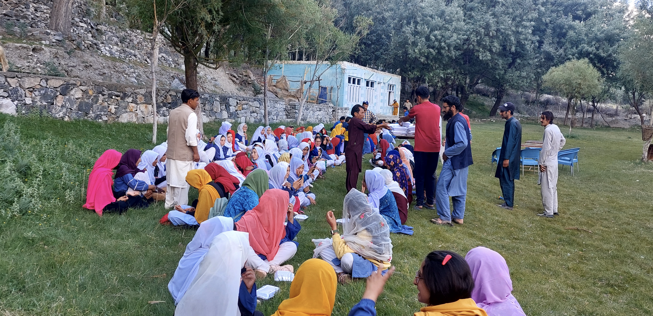 A group of students sitting on the grass in an outdoor setting, with some standing at the front. Most students are wearing headscarves in various colors, and a few adults are distributing papers or talking to the students. The background shows trees, a stone retaining wall, and a small blue building.