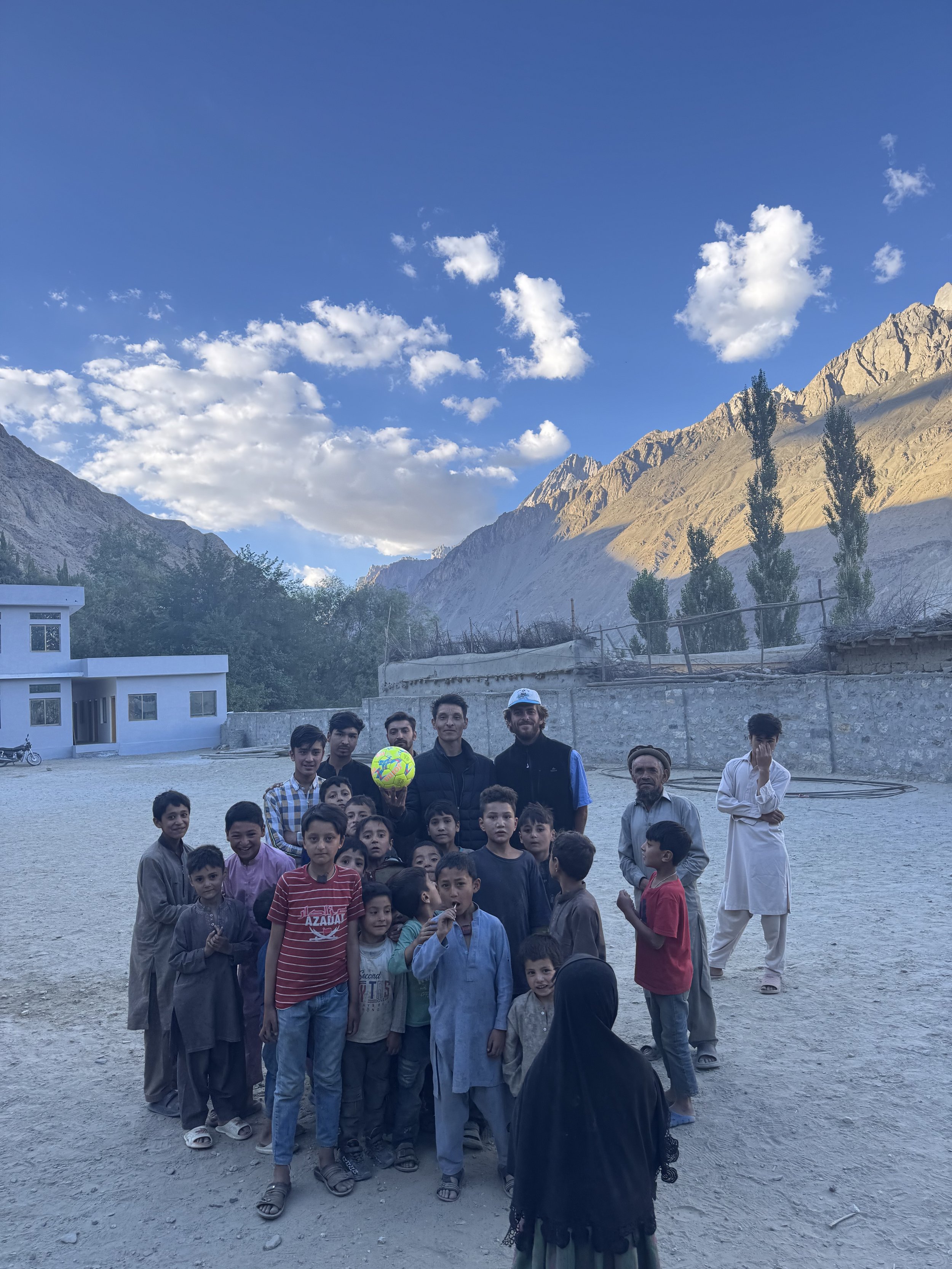Group of children and a few adults in an outdoor setting with mountainous terrain in the background. The children are gathered closely together, some smiling, and one adult is holding a colorful ball. The sky is clear with some clouds.