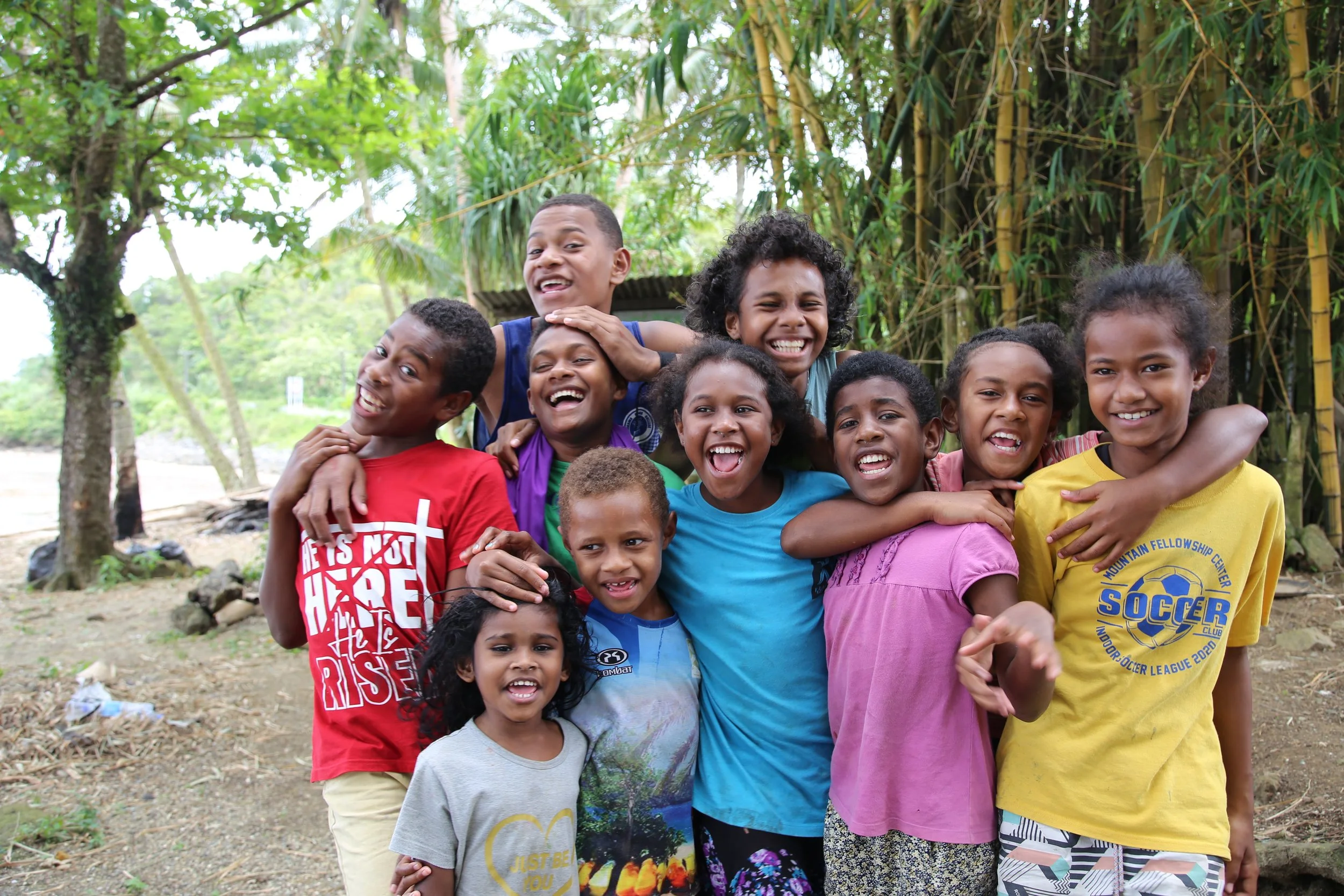 A group of smiling children and teenagers posing outdoors in front of green trees and bamboo, showing camaraderie and happiness.