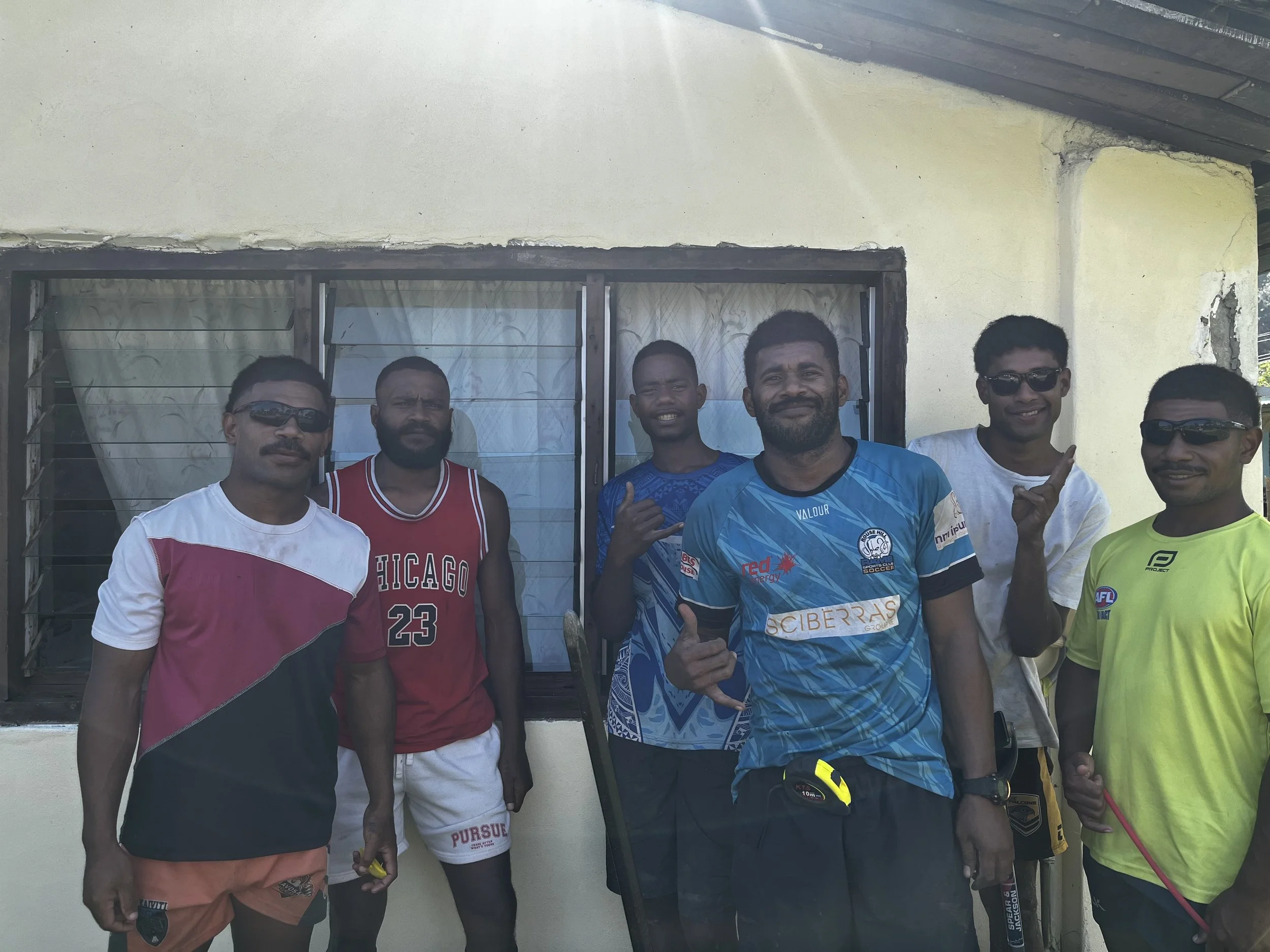 Group of six men standing outside in front of a window with metal bars, some making hand gestures, all casually dressed in sportswear and sunglasses.