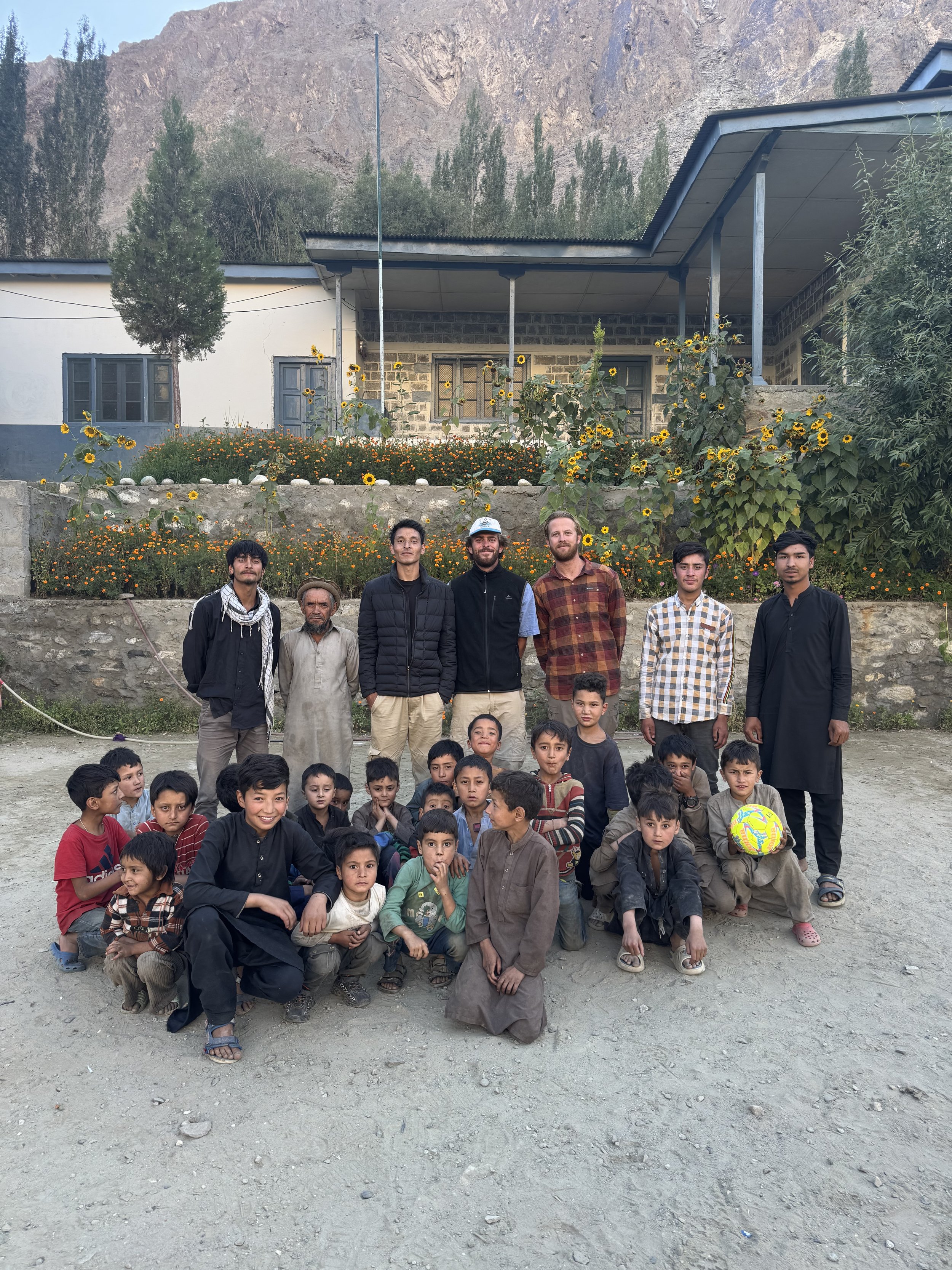 Group photo of adults and children outdoors with a building, flowers, and mountains in the background.