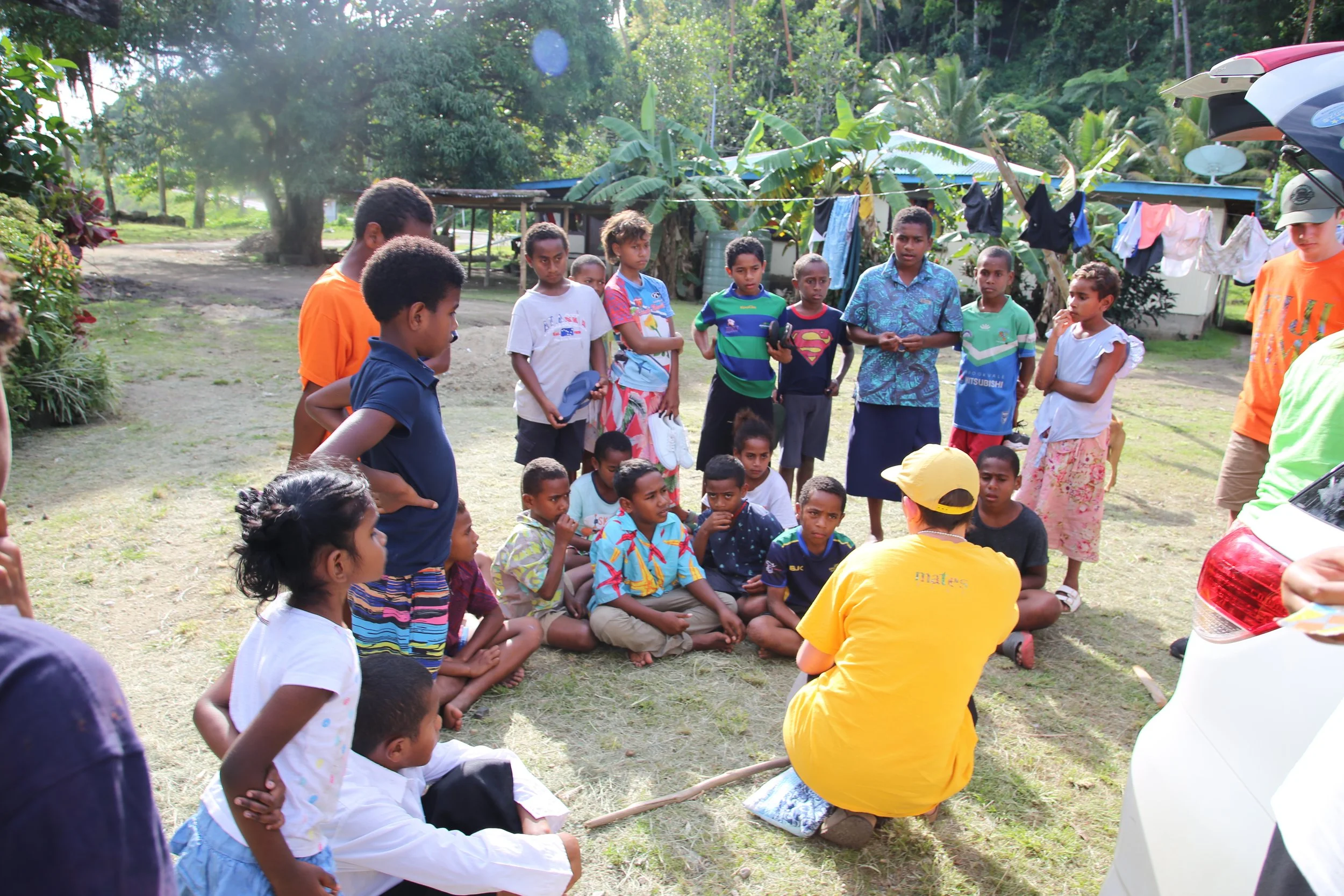 Group of children and adults gathered outdoors, sitting and standing on grass, listening to a person in a yellow shirt and hat who appears to be giving a talk or demonstration. There are trees and makeshift clothing lines with laundry hanging in the background.