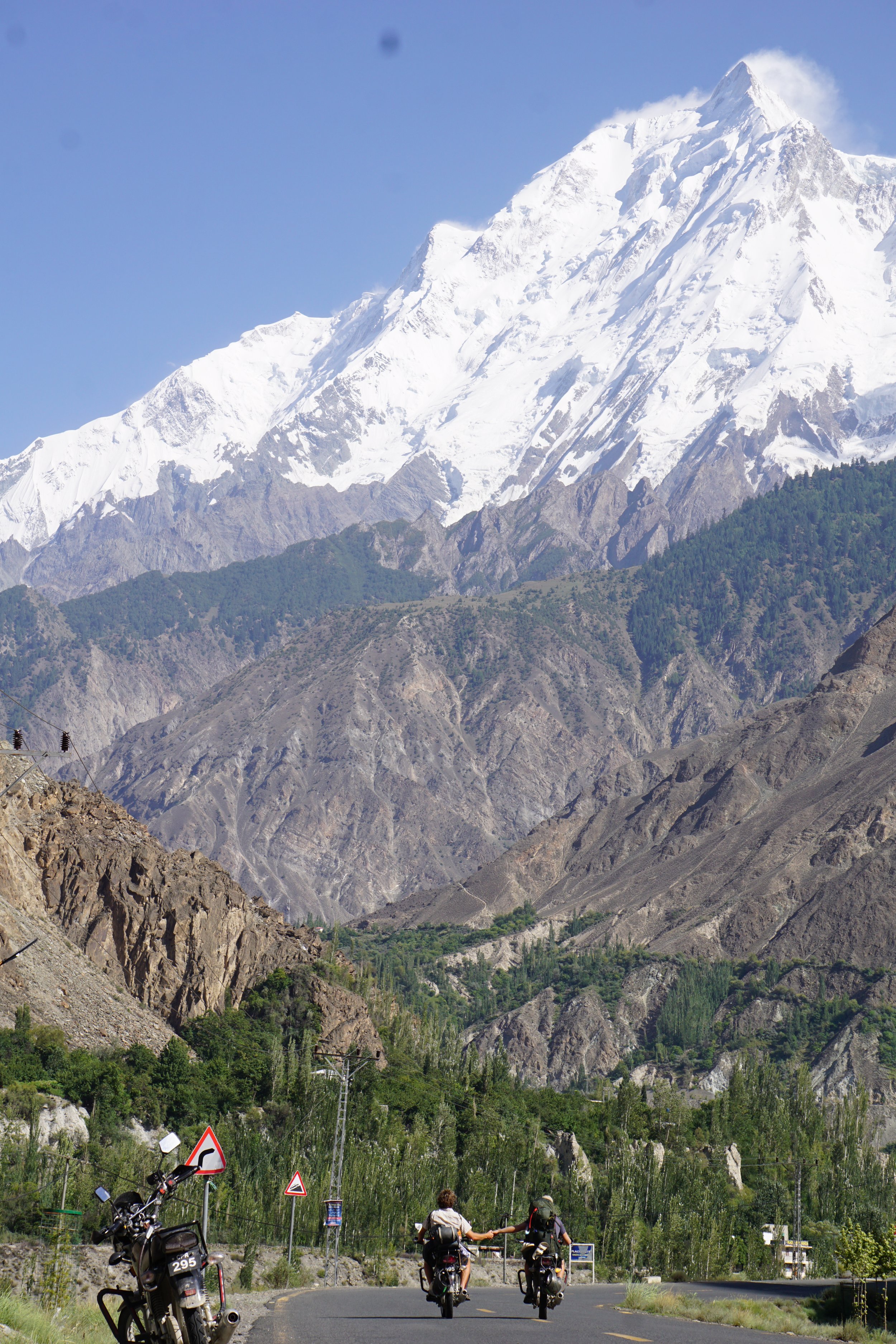 Motorcycles riding on a mountain road with snow-capped mountains and green trees in the background.