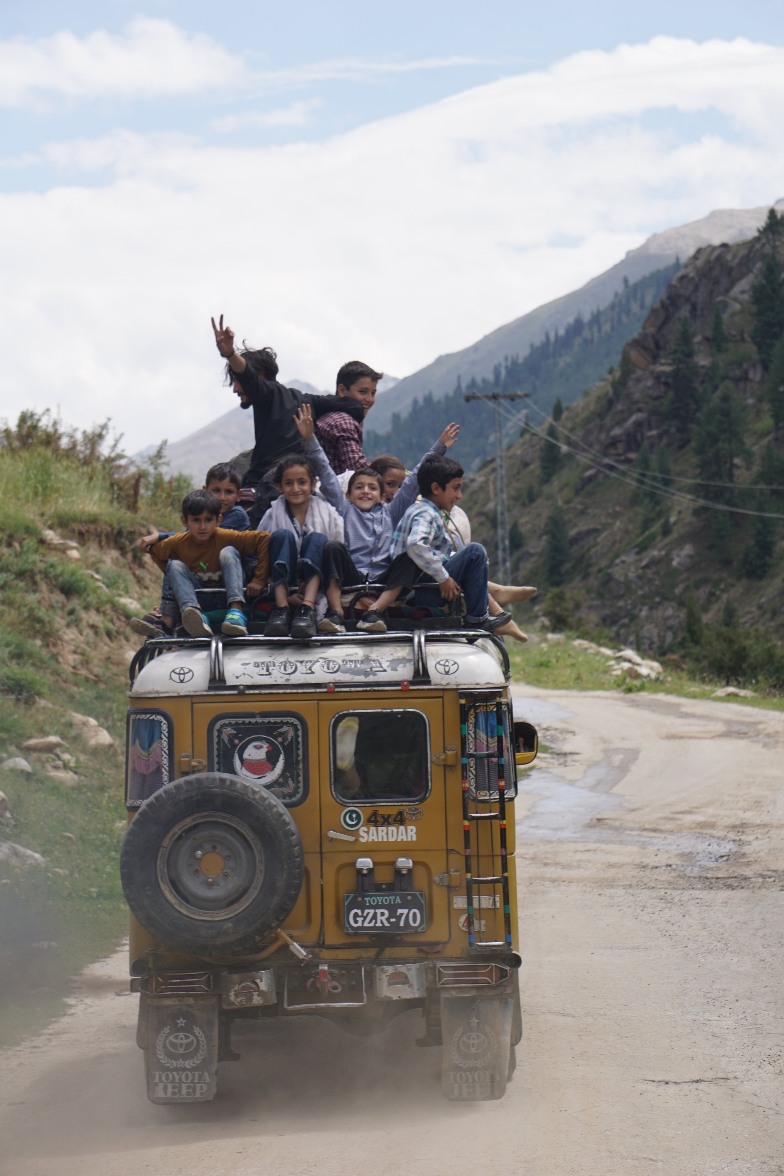 Children riding on top of a yellow Toyota jeep on a dirt road in a mountainous area, some waving and smiling.