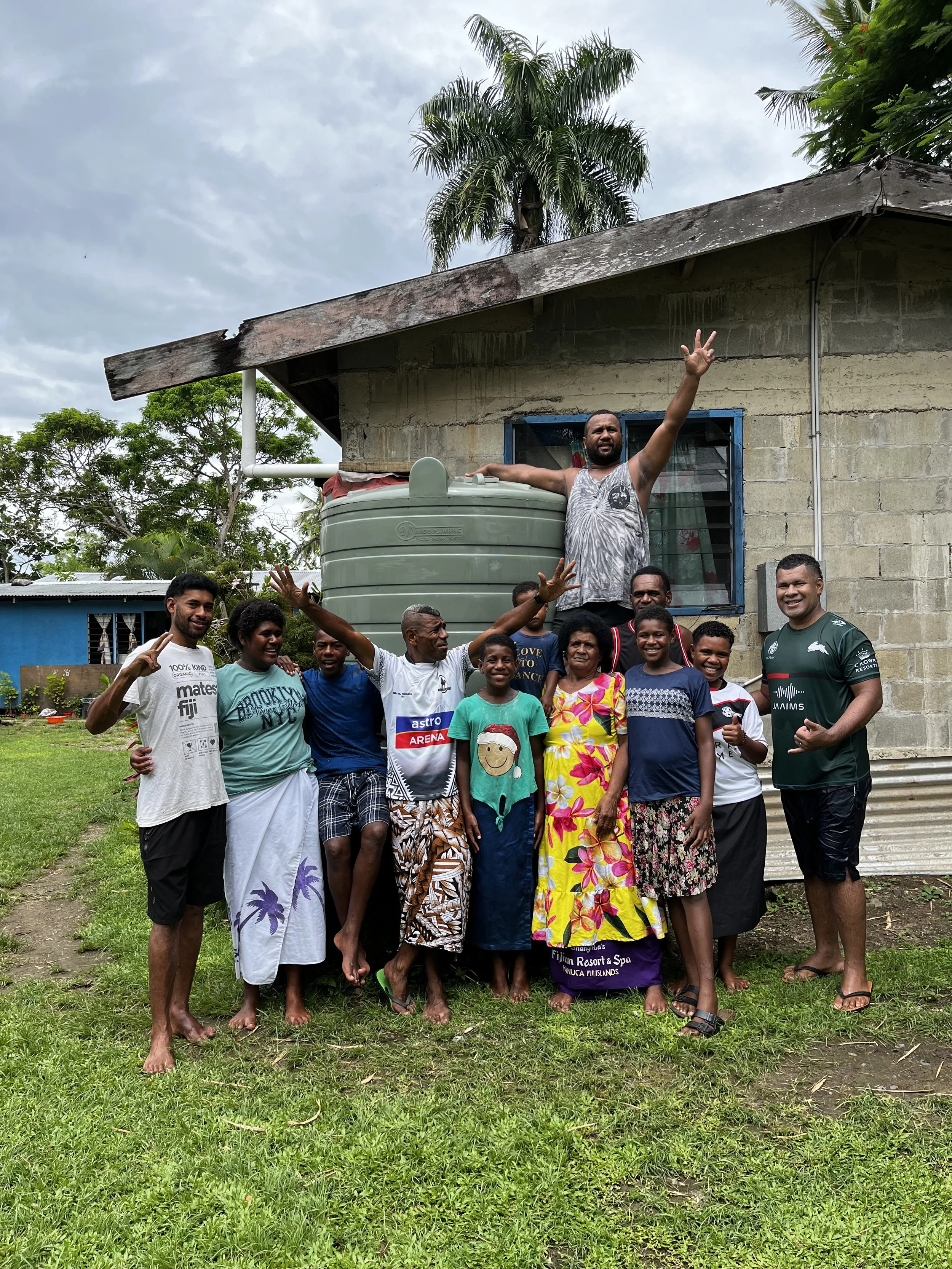 Group of people standing together outdoors in front of a house with a green water tank, some smiling and waving, dressed casually, with a cloudy sky and palm trees in the background.