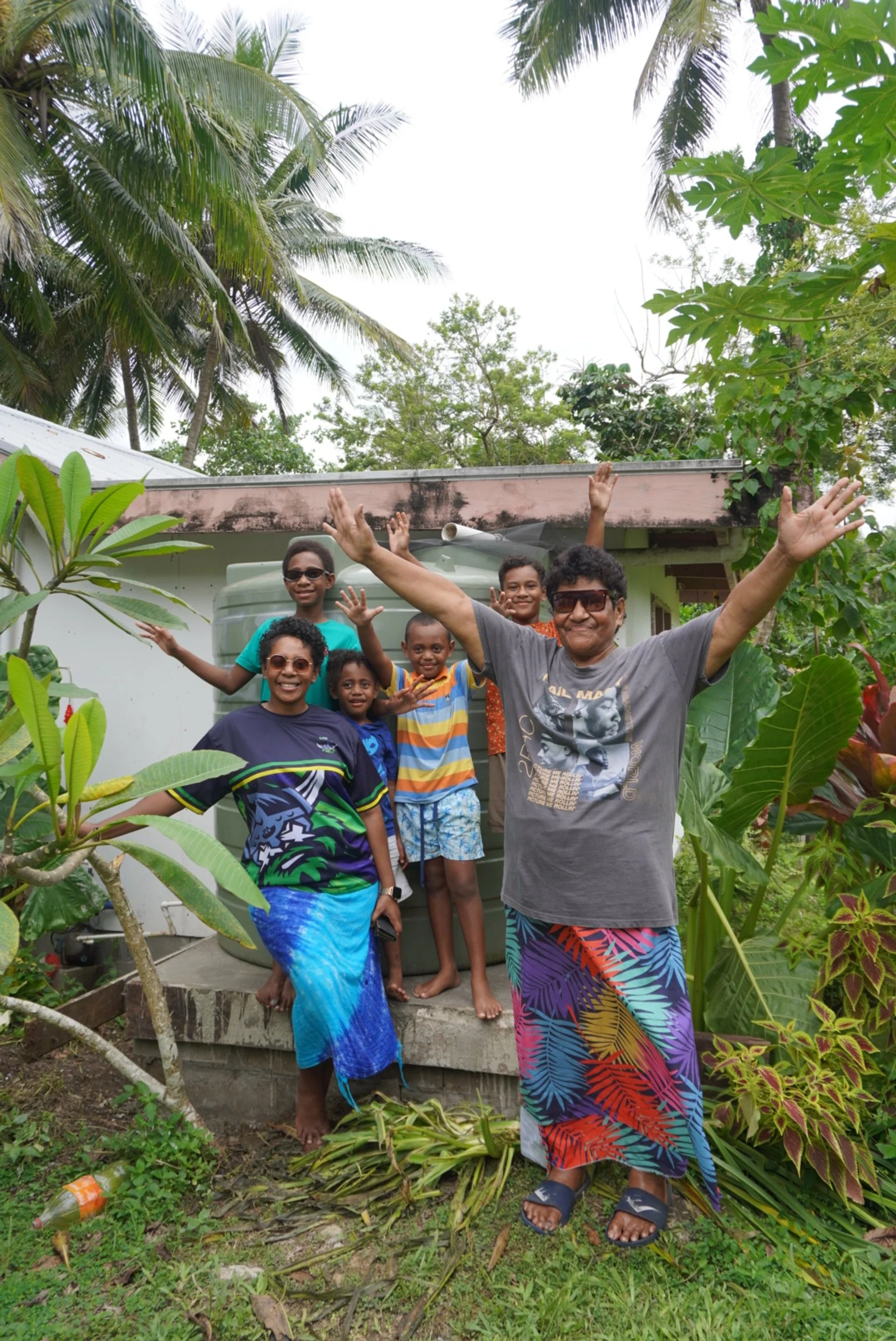 A group of six people, including one adult and five children, standing outdoors in a tropical garden with lush greenery and palm trees, smiling and raising their hands in celebration.