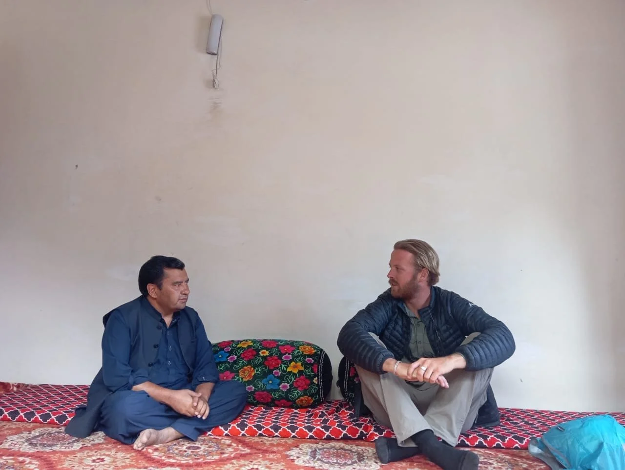 Two men are seated on a decorated carpeted floor against a plain off-white wall, engaged in conversation. The man on the left is wearing a dark blue traditional outfit, and the man on the right is wearing a dark jacket over a shirt and light-colored pants, with a backpack on the floor nearby.