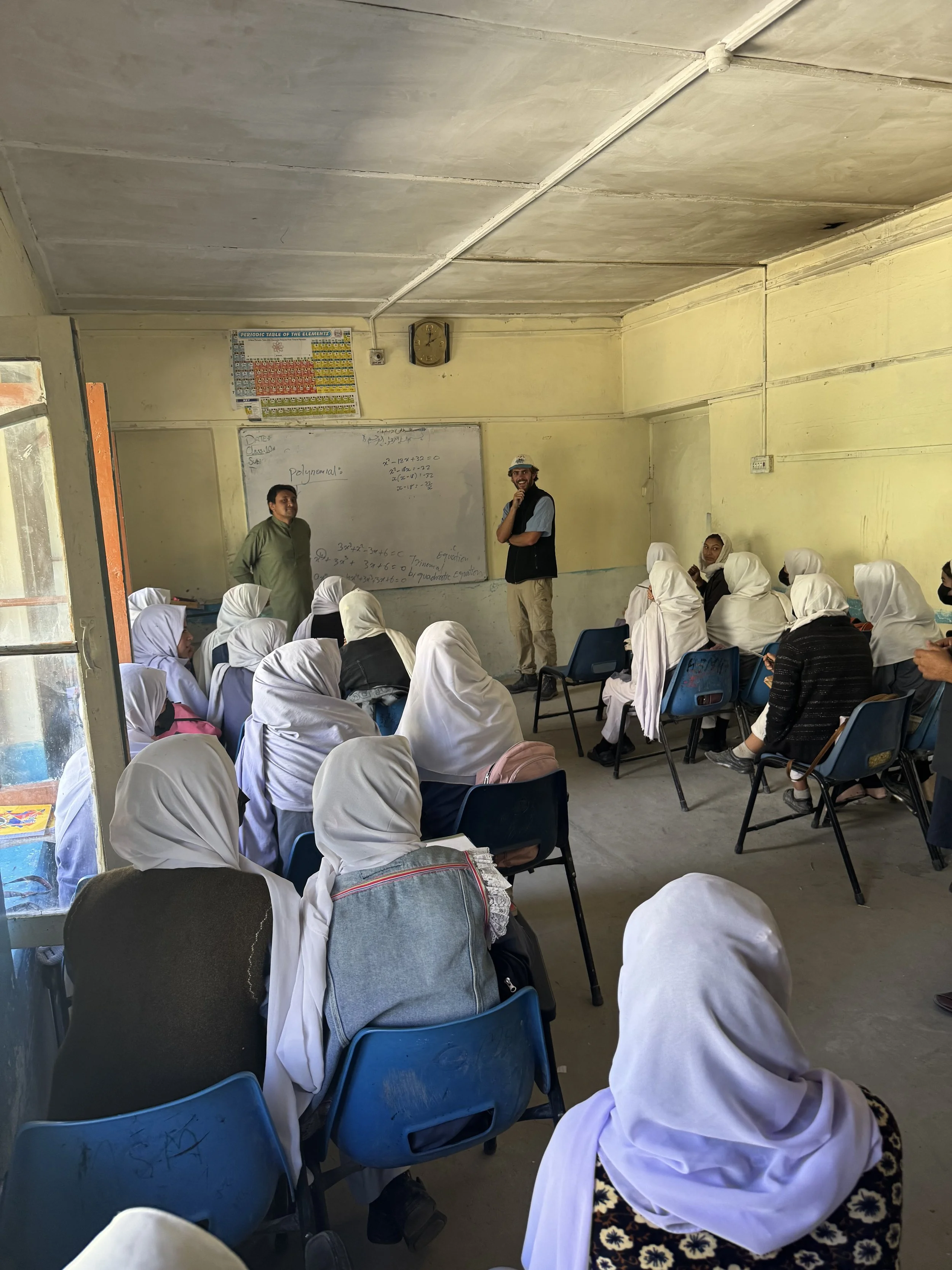 A classroom filled with students wearing white headscarves, listening to two male teachers at the front. One teacher is standing near the whiteboard, the other is smiling and covering his mouth, both engaged in teaching.