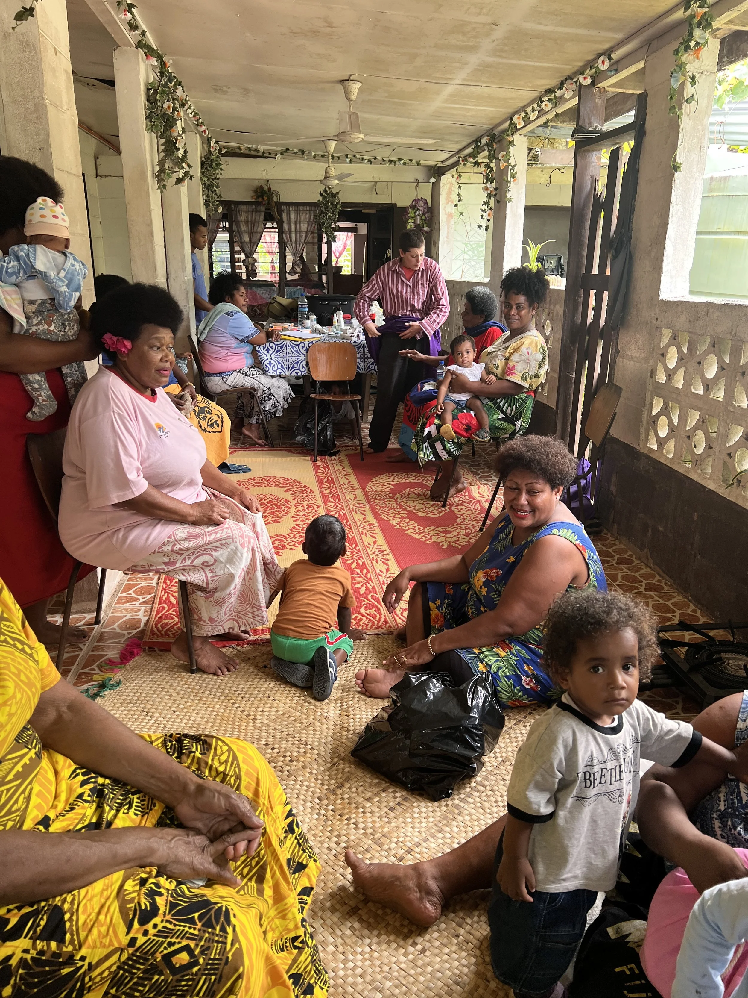Group of people in a cozy, decorated indoor space with hanging plants, sitting on chairs and mats, some with children, engaging in a social gathering.
