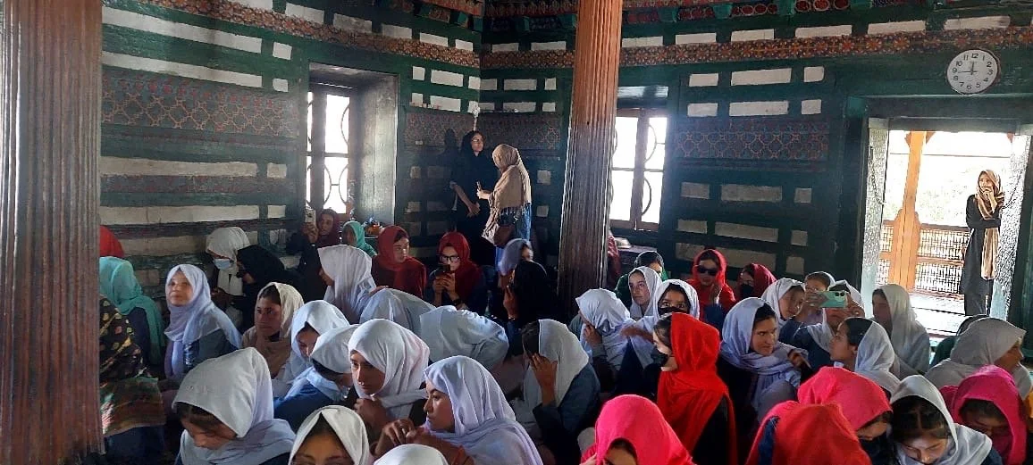 A group of women and girls sitting inside a traditional wooden building with patterned walls, some wearing headscarves, while a few are standing and talking.