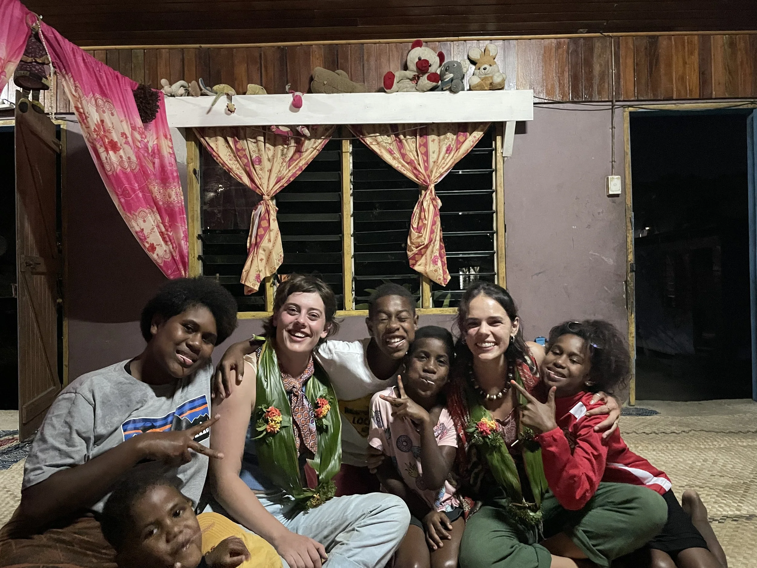 Group of six smiling people, including children, sitting together inside a room with wooden walls and pink curtains, some wearing flower garlands, posing for a photo.