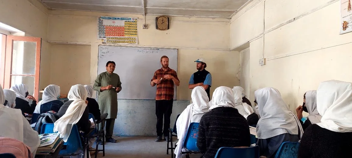 Classroom scene with students wearing white headscarves seated at desks listening to three male teachers at the front, with a whiteboard and world map on the wall.