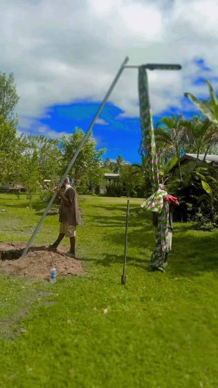People installing a tall metal pole in a grassy yard on a cloudy day, with trees and houses in the background.
