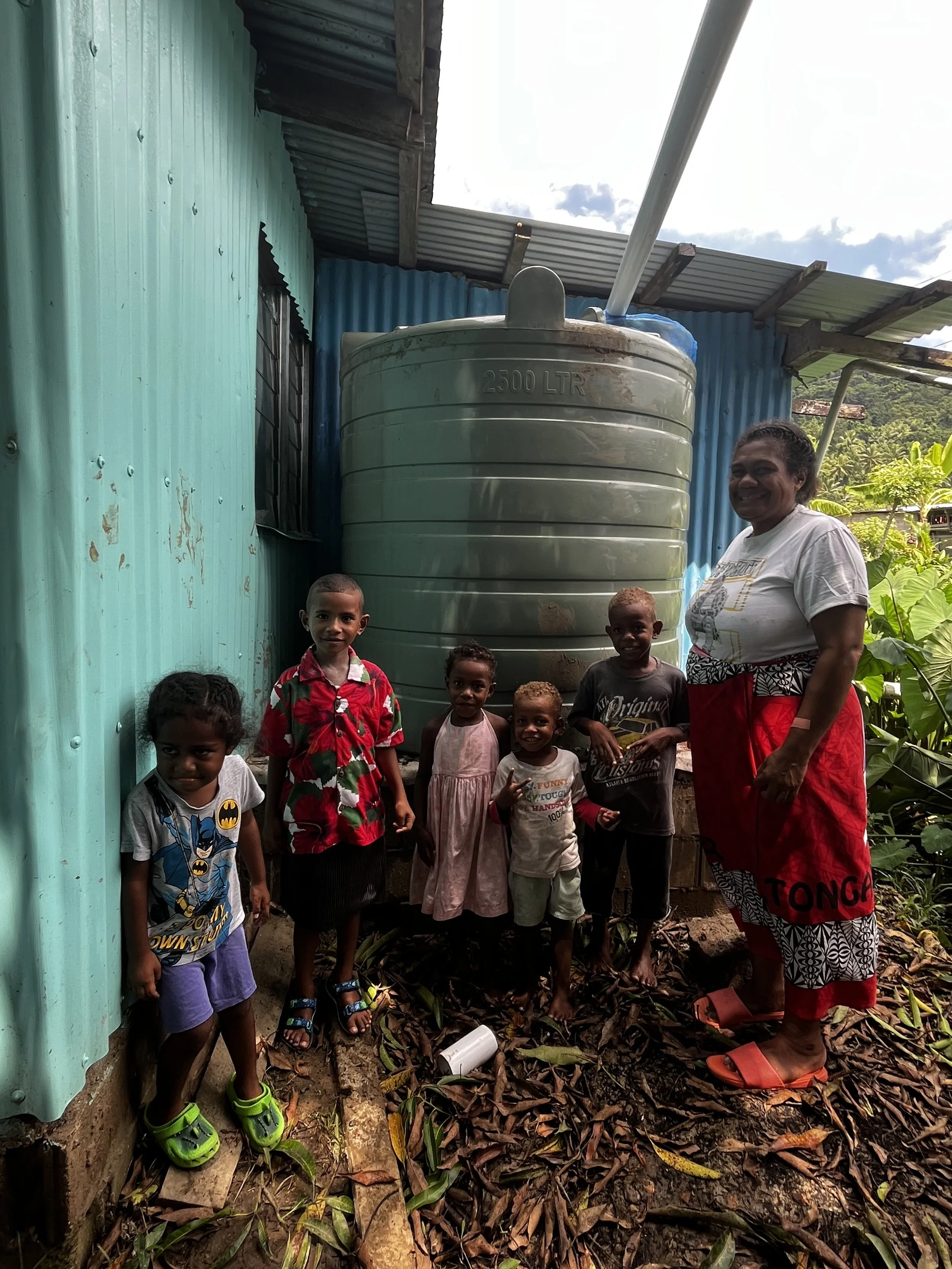 A woman and six children standing outside next to a large 2500-liter water tank, with a corrugated metal wall and lush greenery in the background.