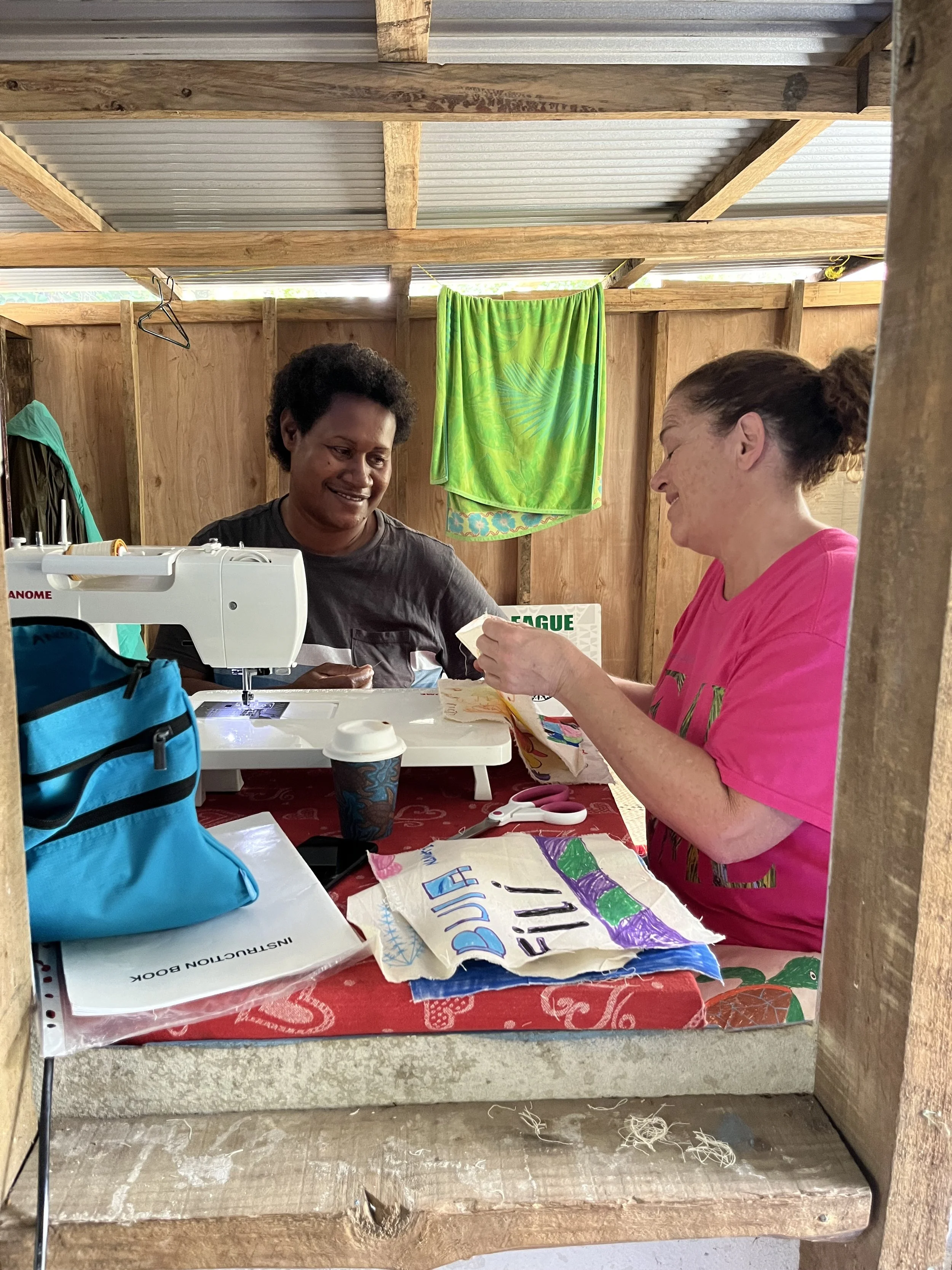 Two women are sitting at a table inside a wooden shelter. One woman is sewing on a sewing machine, while the other woman is handing her a piece of fabric. The table has various sewing supplies, including fabric pieces with colorful designs, scissors, a coffee cup, and an instructional book. A green towel hangs on the wall, and the shelter has a corrugated metal roof.