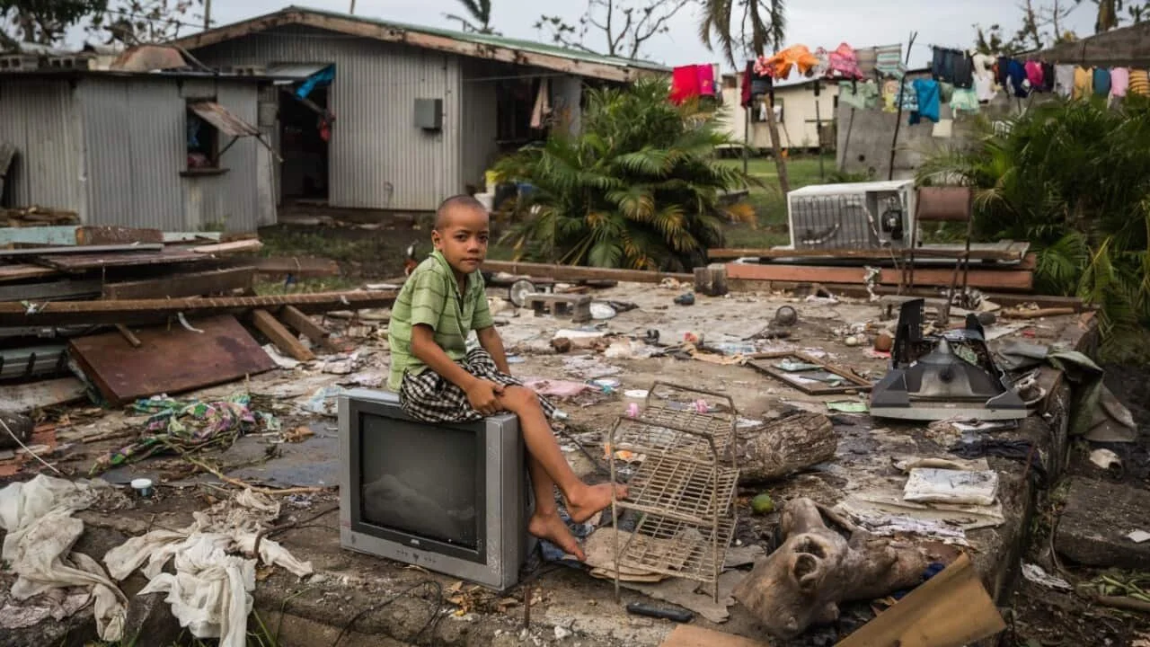 A young boy sits on an old television amidst debris and trash in a damaged or destroyed outdoor area, with damaged buildings and laundry hanging in the background.