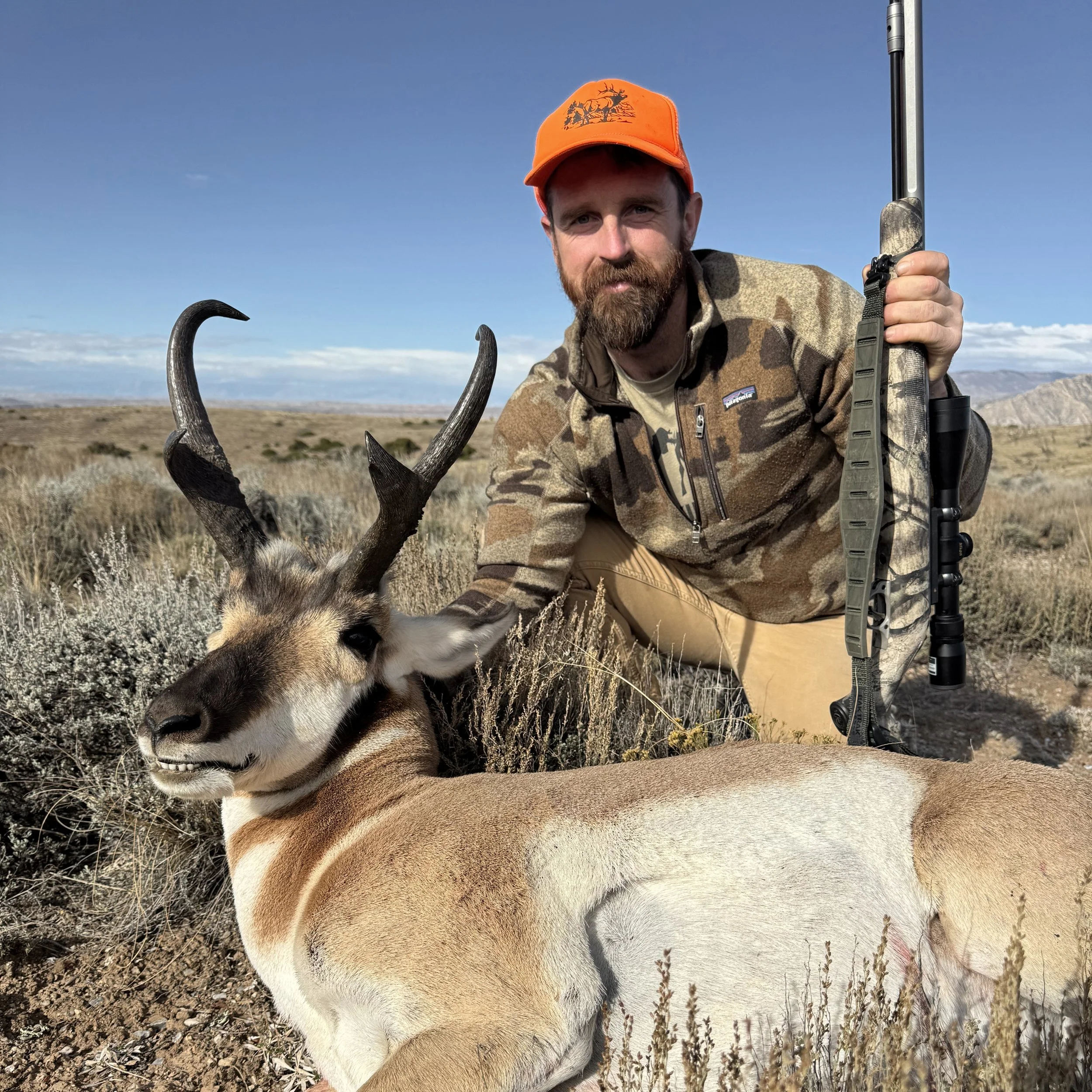 A man with a beard wearing an orange hat and camouflage jacket kneeling next to a harvested antelope with large curved horns in a grassy open field with mountains in the background.