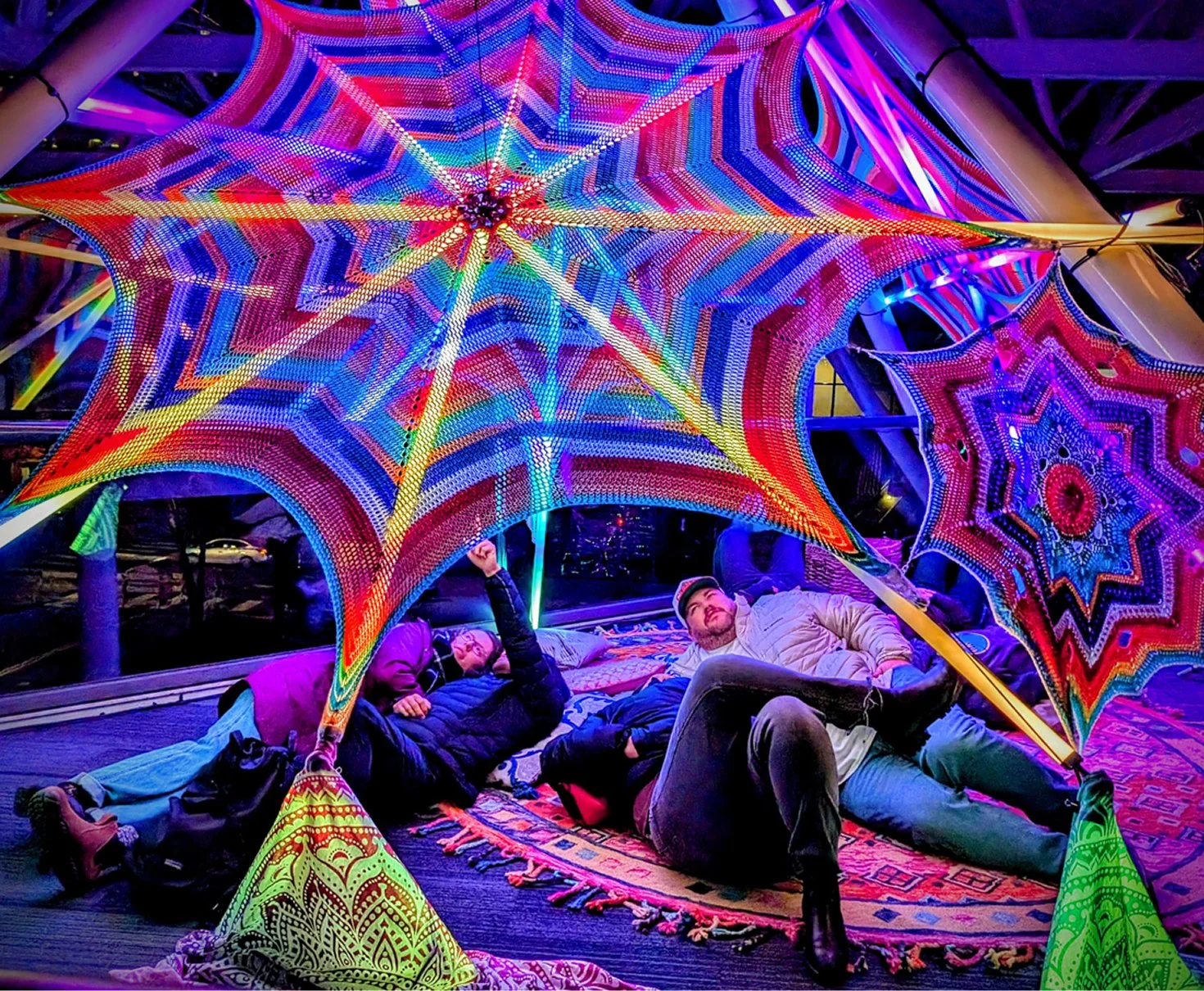 Colorful, illuminated, mandala-shaped decorations resembling spider webs, with people lying and sitting underneath, inside a modern interior space.