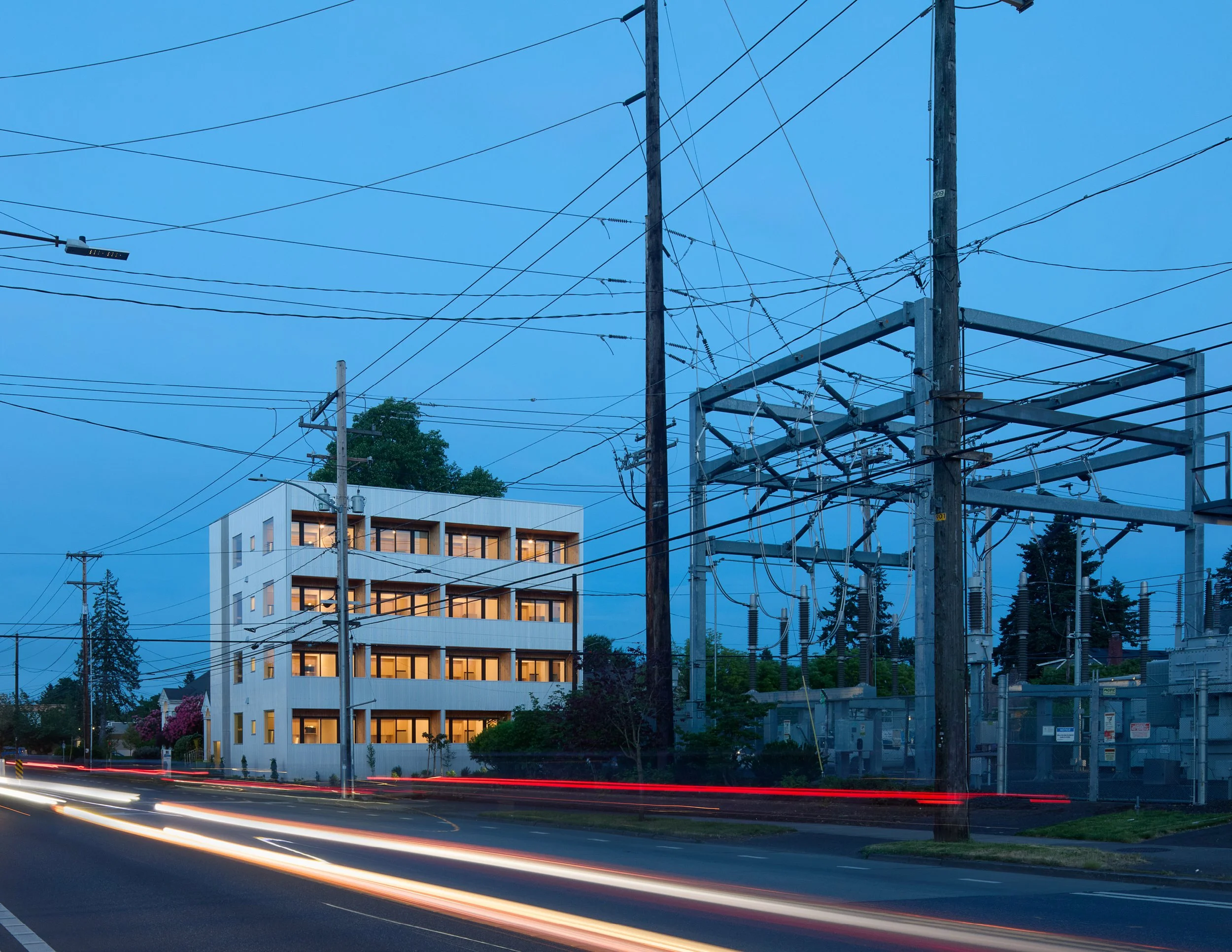 Modern two-story house with a garage and large glass windows, illuminated interior at dusk, surrounded by trees.